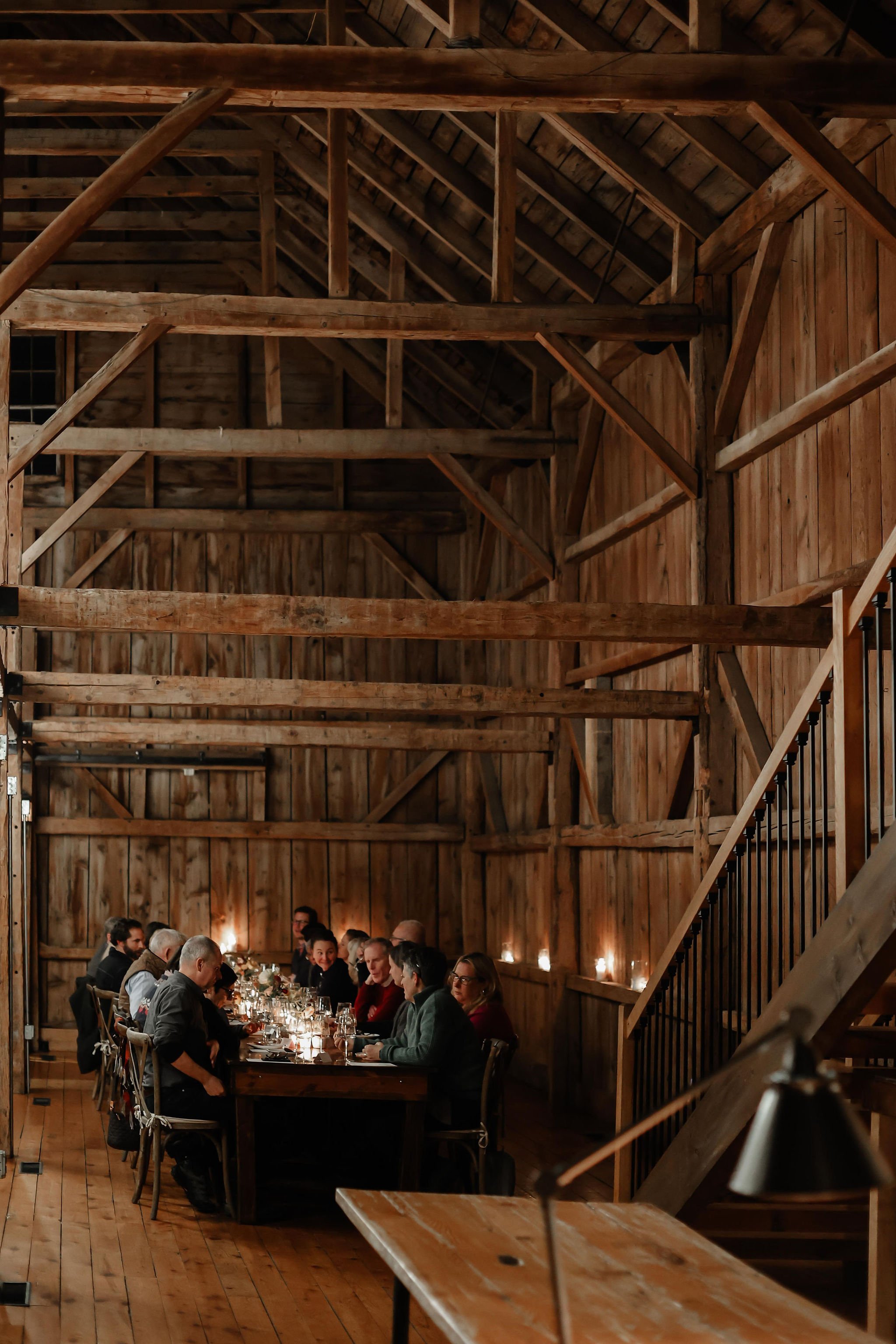 A group of people gathered around a long dinner table in a rustic wooden barn.