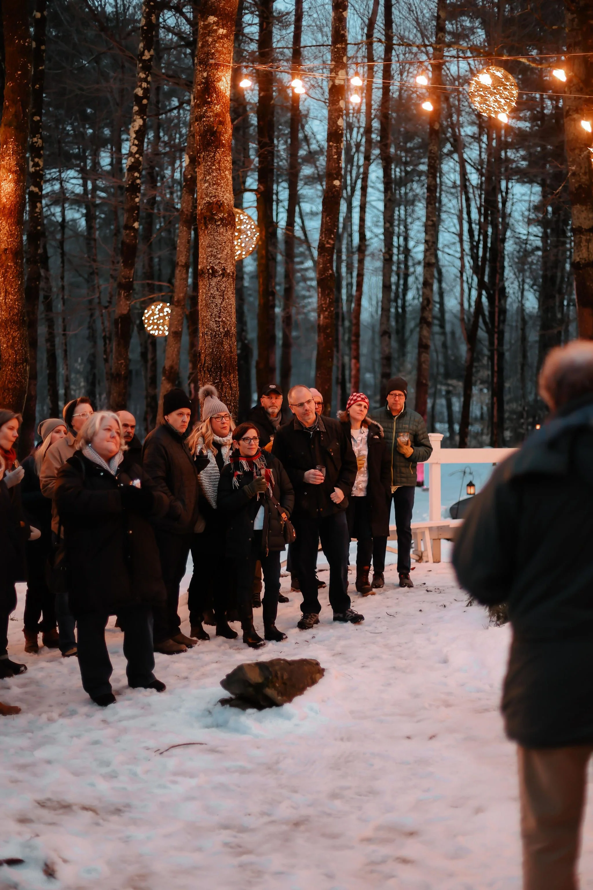 A group of people gathered outdoors in a snowy wooded area, watching something or listening to a speaker, with string lights hanging among the trees.
