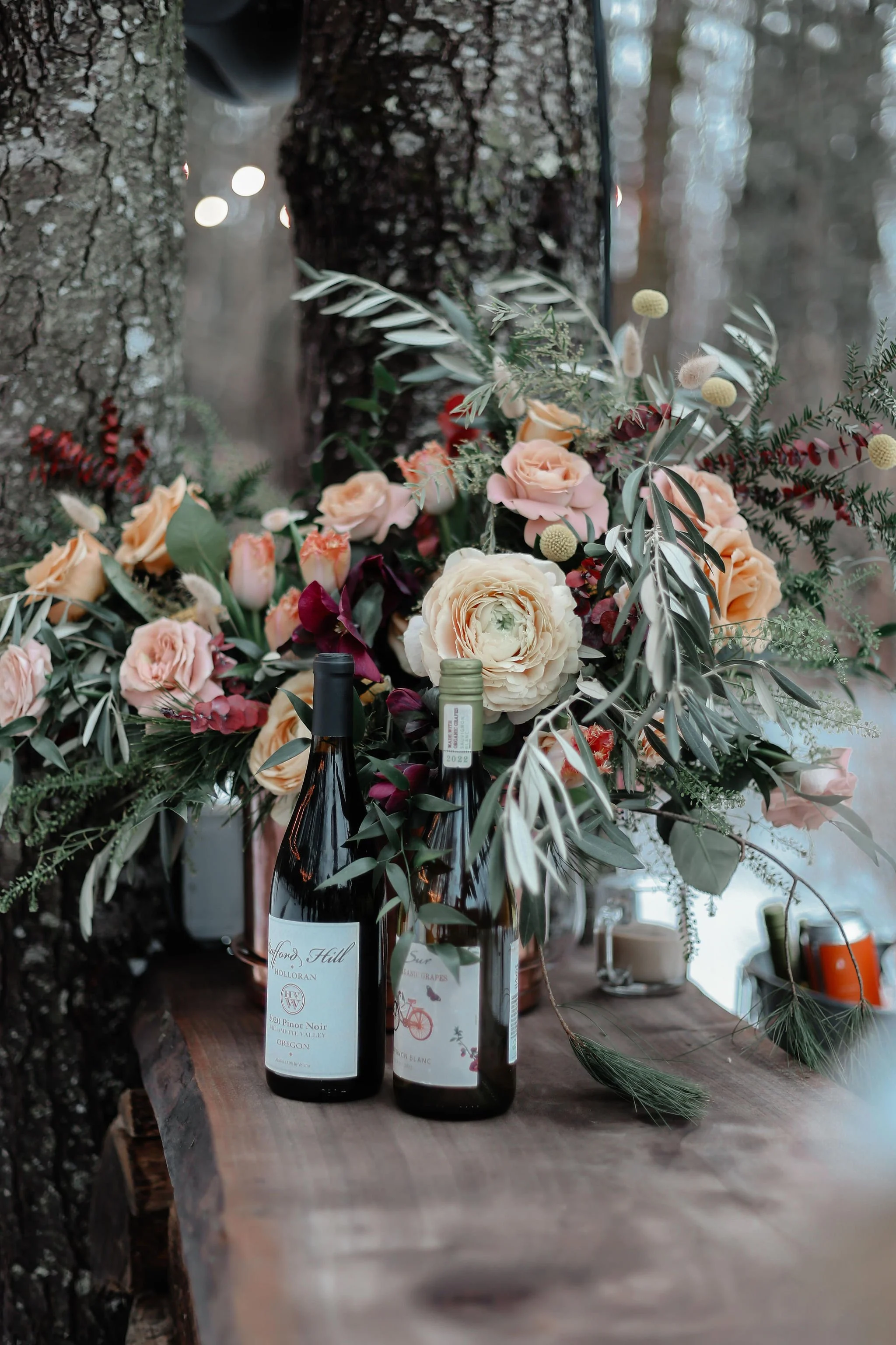 A floral arrangement in bottles and vases with peach and burgundy flowers and greenery, placed on a wooden table outdoors, against a backdrop of trees and string lights.