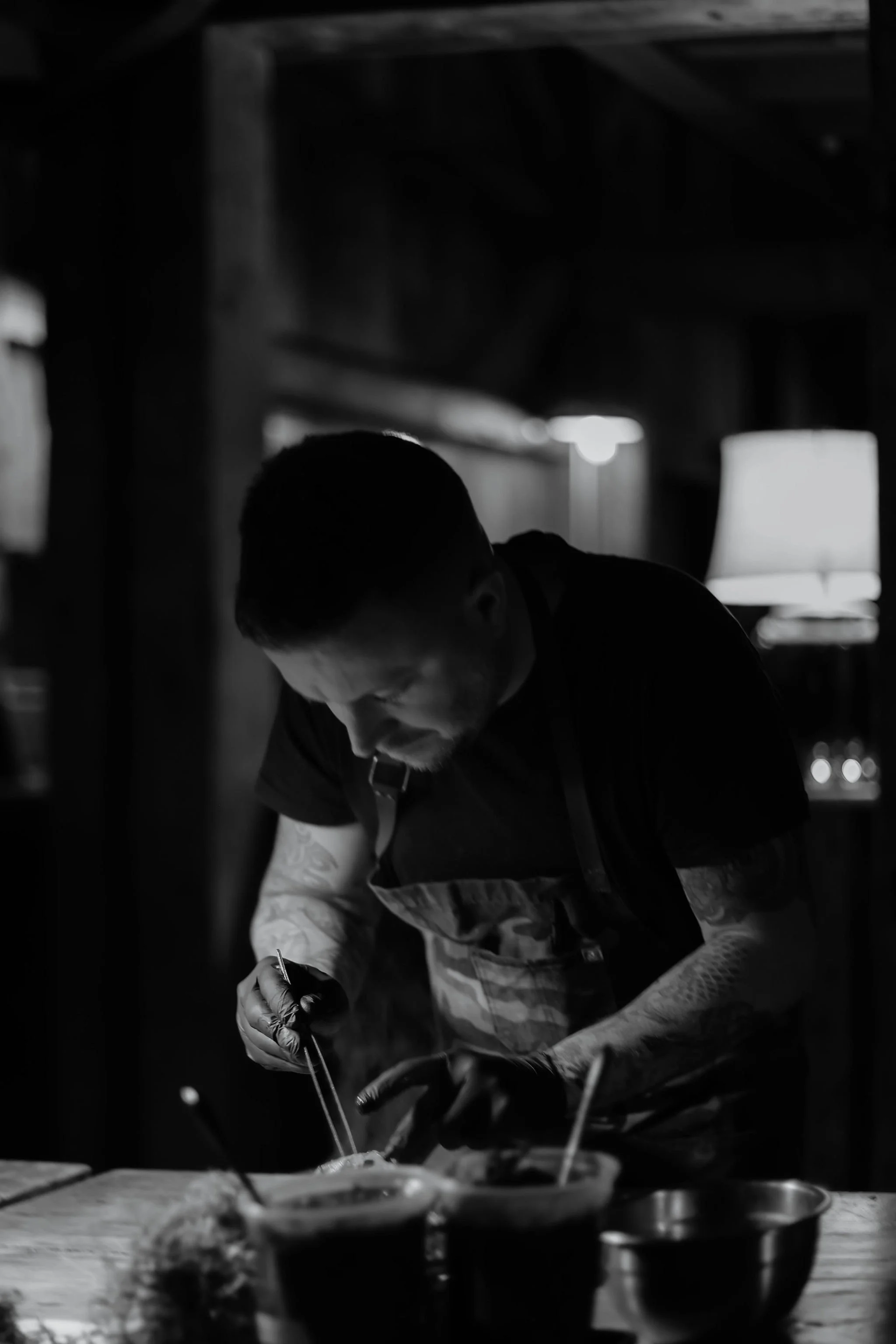 A man prepares food in a dimly lit, cozy room, wearing an apron and working intently over a table with various bowls.