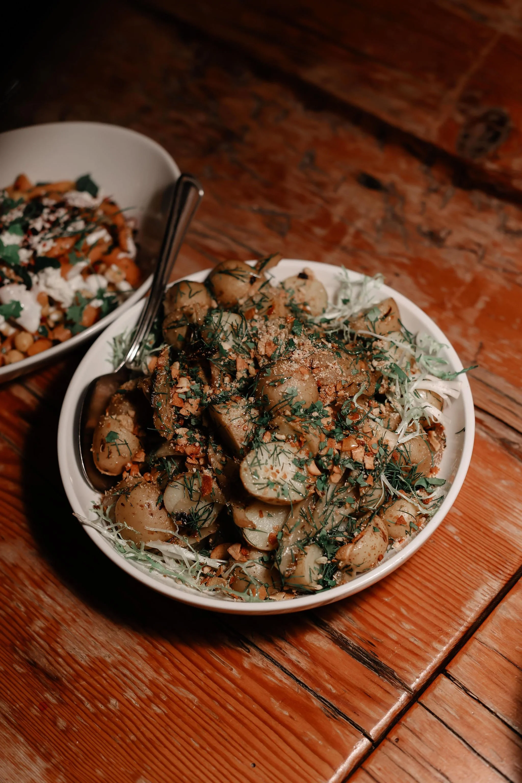 A white bowl filled with marinated mushrooms topped with herbs and crushed nuts, placed on a wooden table. There is another bowl with a salad in the background.