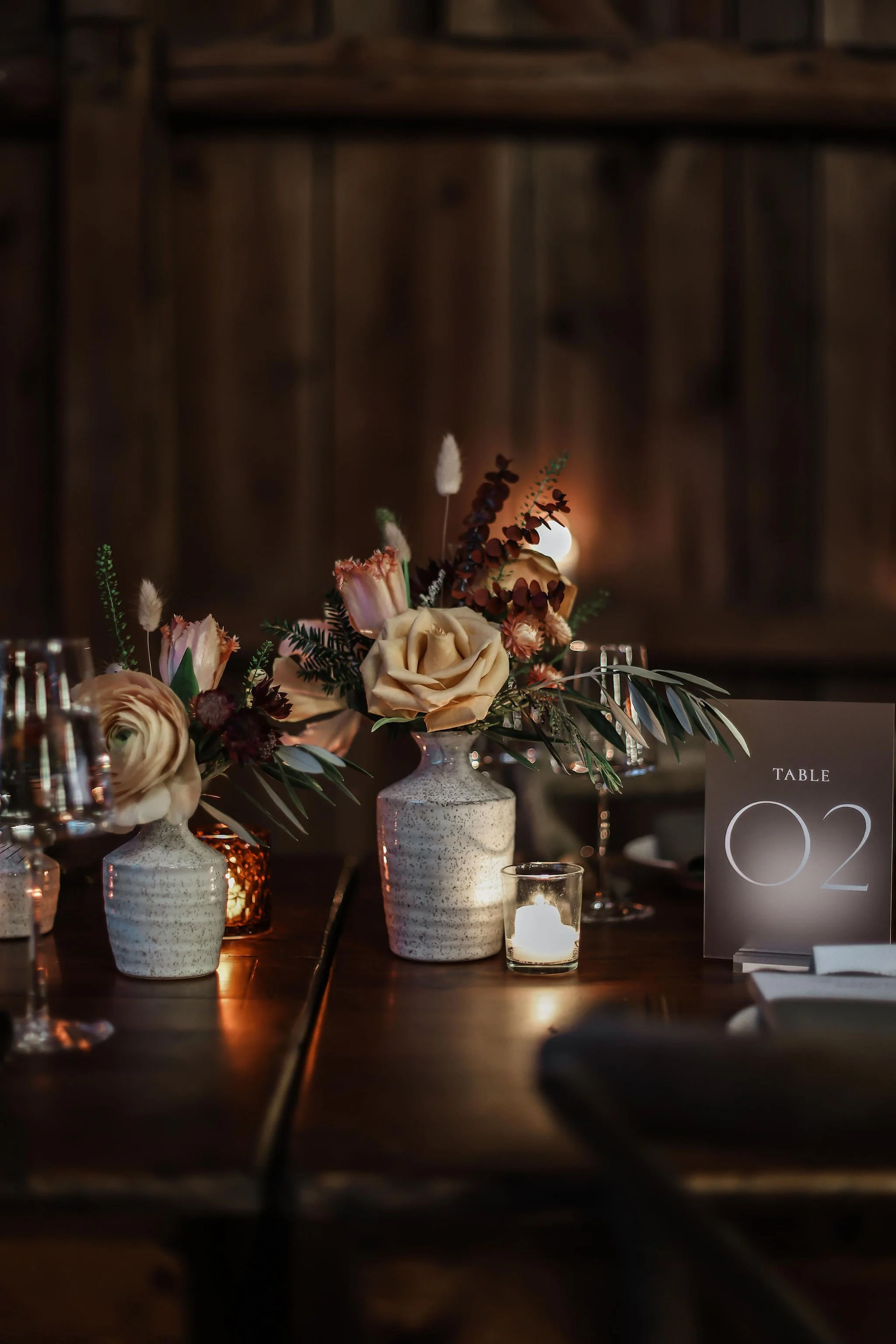 A floral centerpiece with cream and pink flowers, greenery, and candles on a wooden table in a dimly lit setting, with a table number sign reading 'Table 02'.