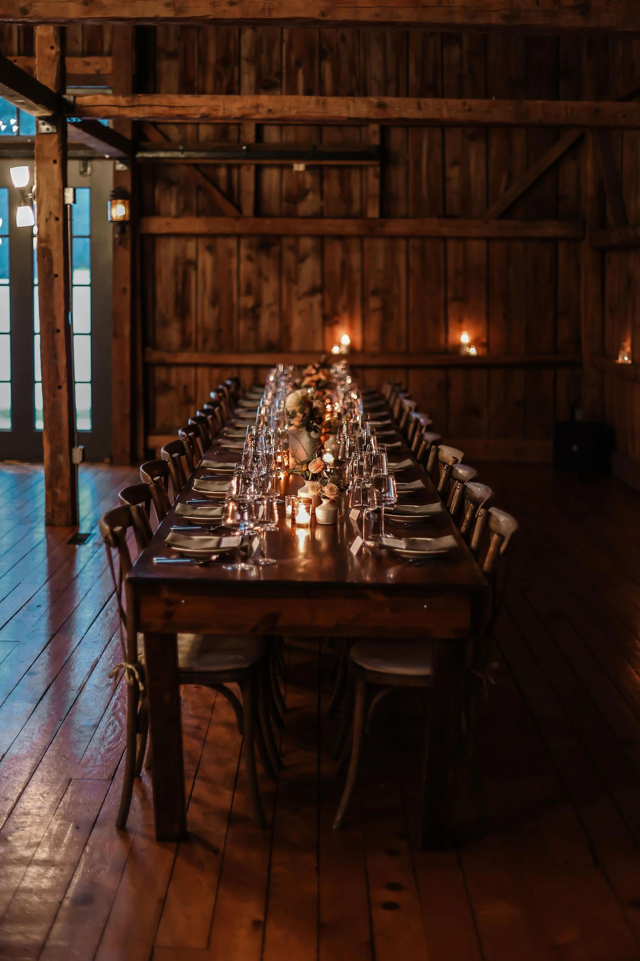 Elegant wooden banquet table decorated with candles, flowers, and glassware in a rustic, dimly lit barn setting.