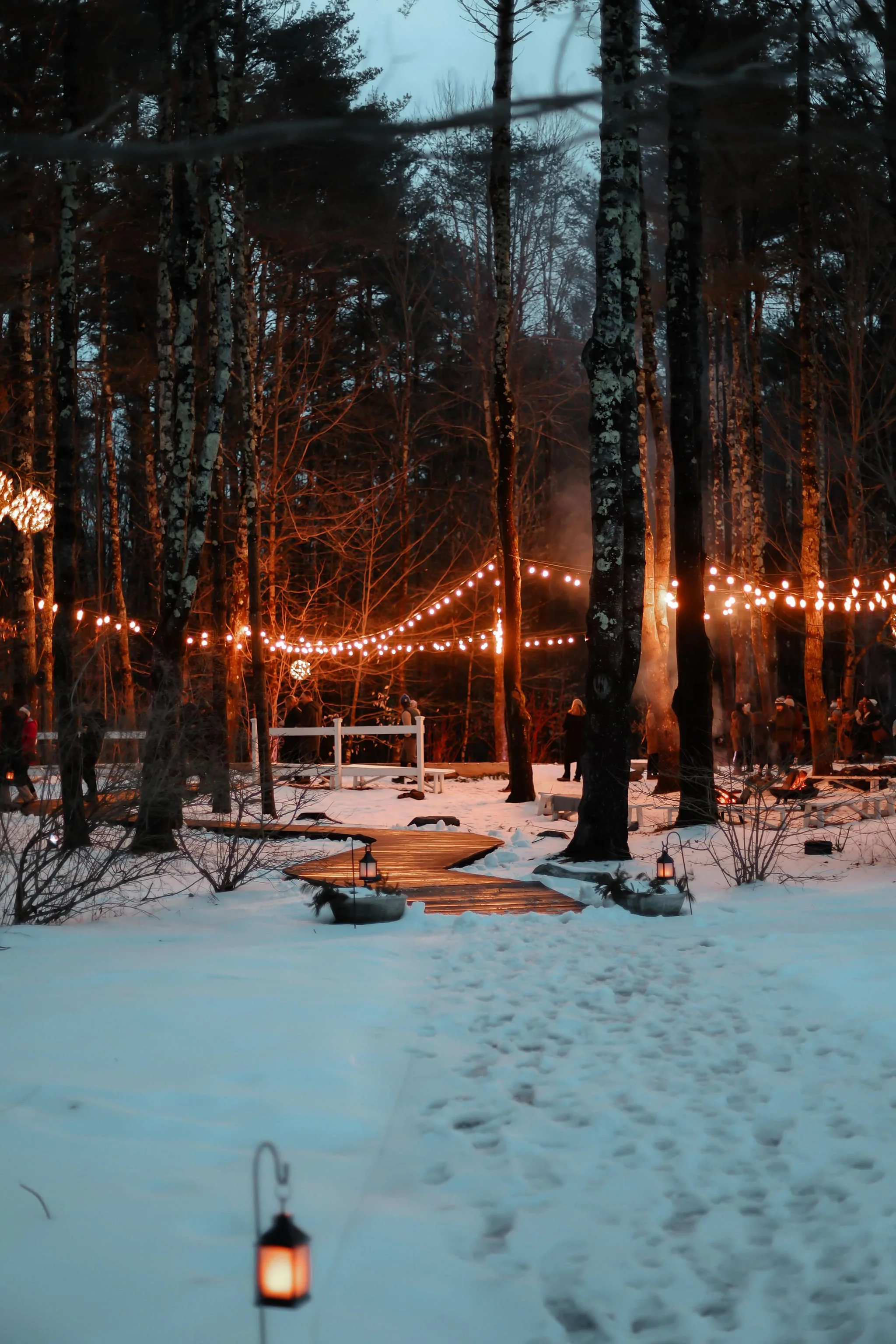 A snowy outdoor scene at dusk with string lights hanging between trees, lanterns on the snow, and a wooden pathway leading through the woods, with people gathered in the background.