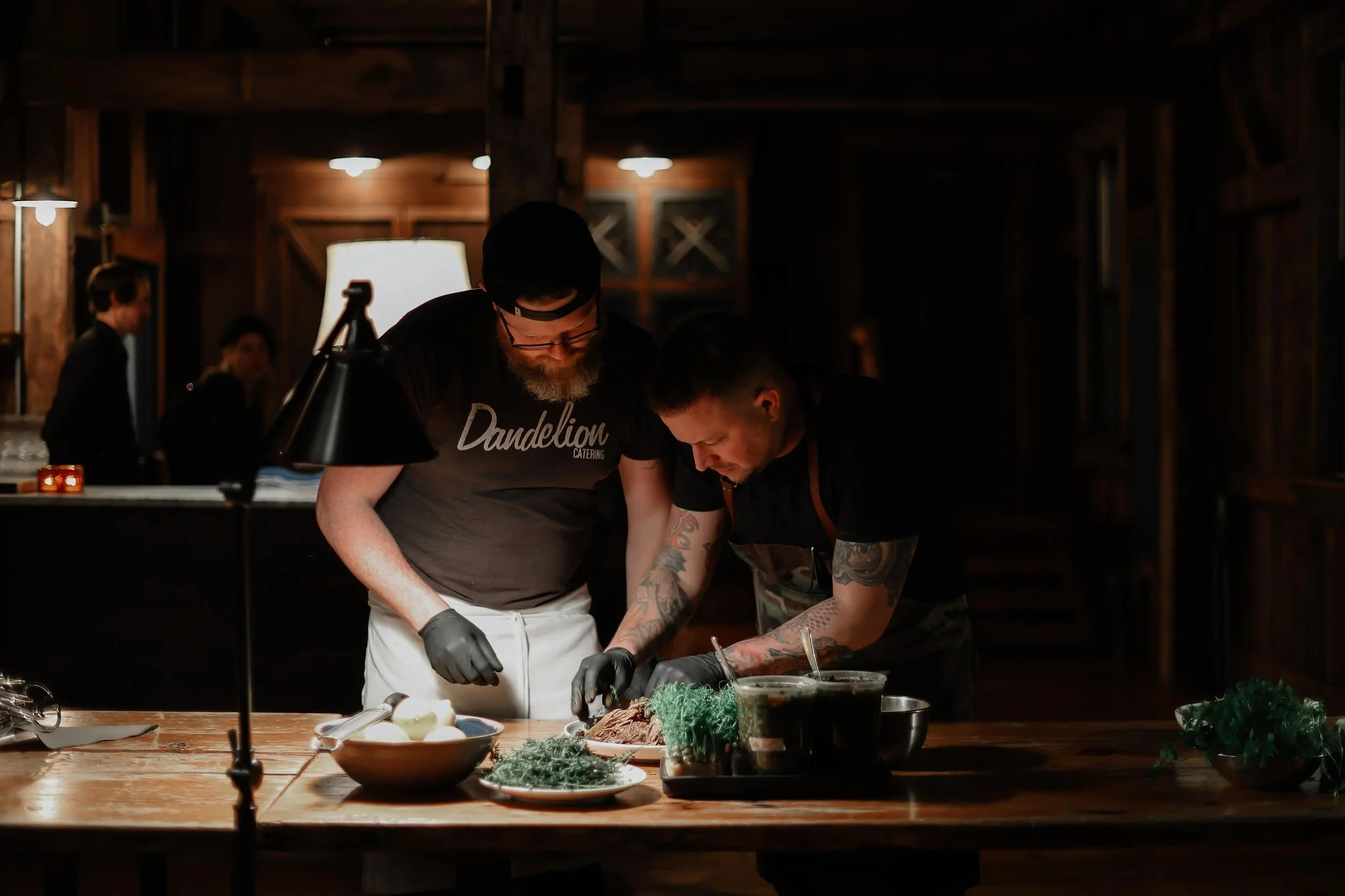 Two men in black T-shirts and gloves preparing food on a wooden table in a rustic, dimly lit kitchen or restaurant, with additional staff working in the background.