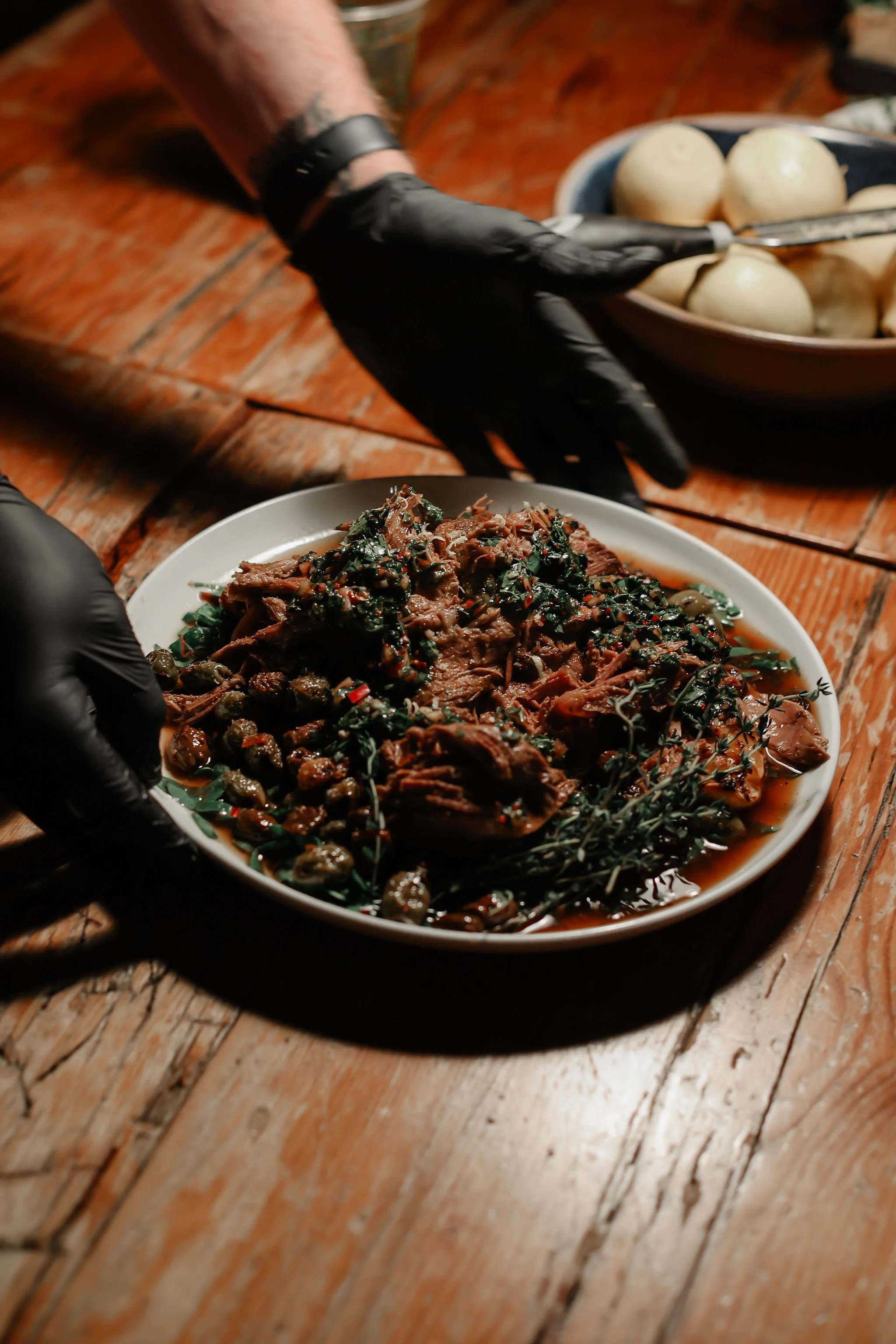 Close-up of a plate of cooked meat and herbs, with a person's gloved hand nearby, on a rustic wooden table.