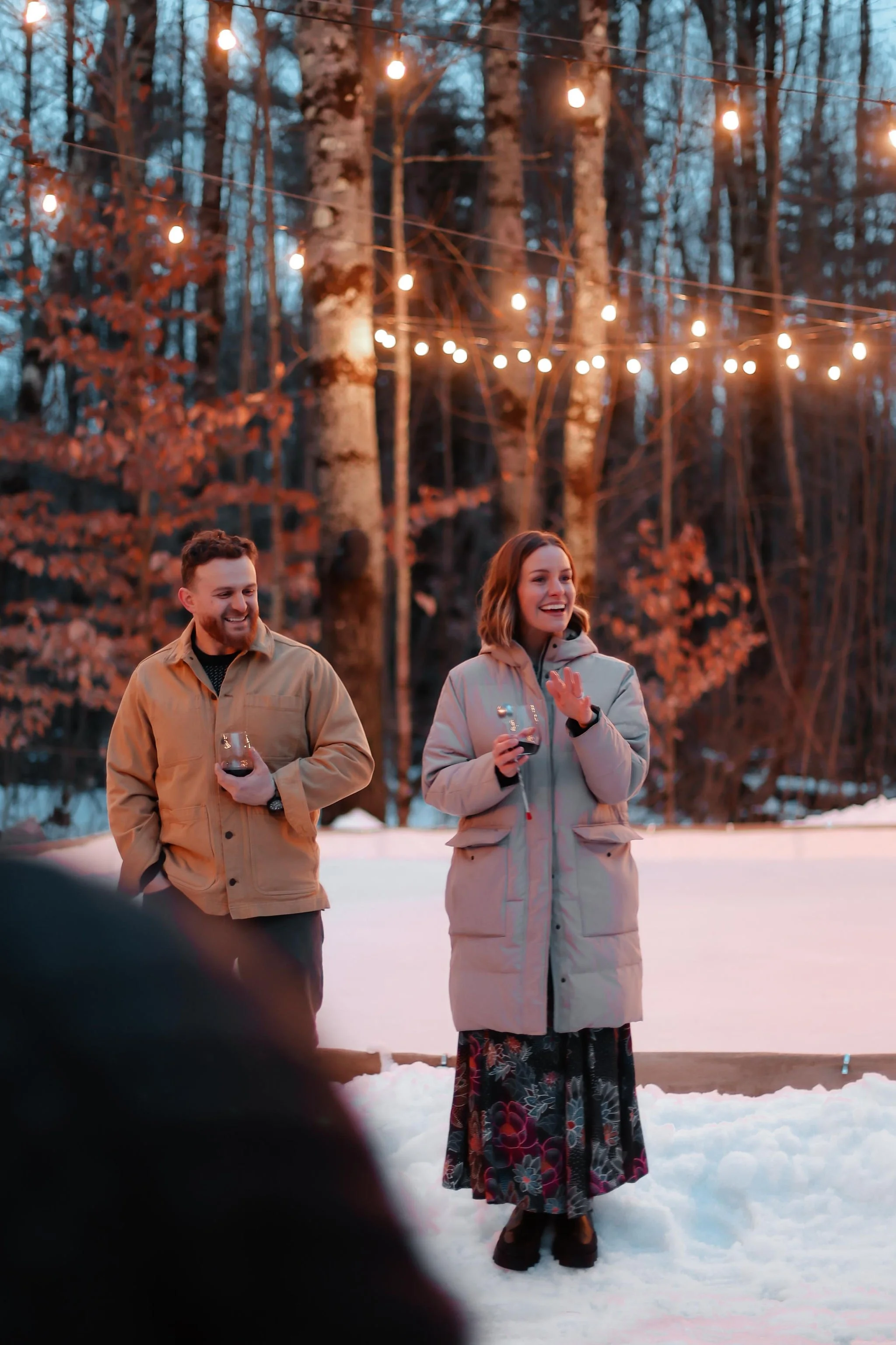 A man and a woman stand outside in the snow, holding glasses of red wine, laughing and enjoying a social gathering. String lights hang above them in a wooded area, creating a warm ambiance at dusk or evening.
