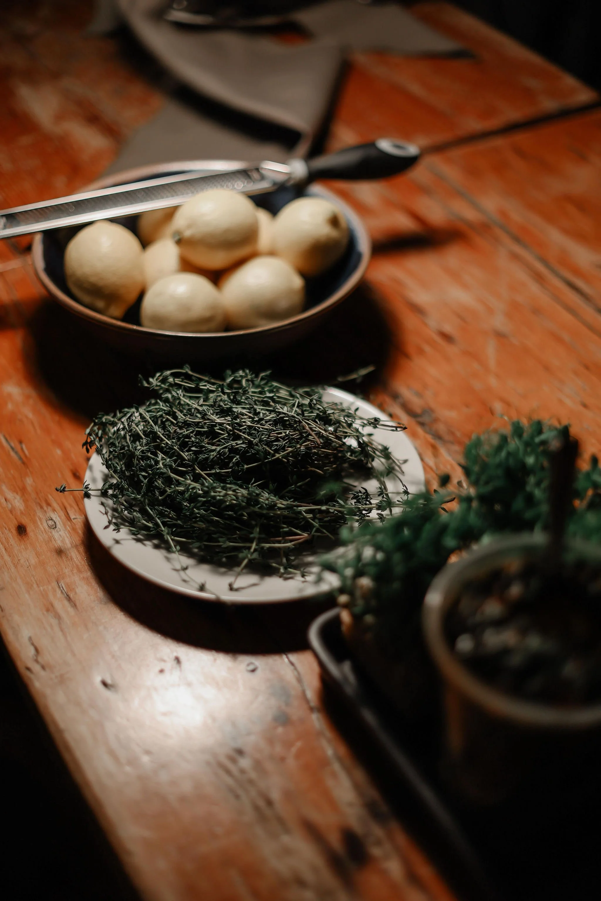 A wooden table with a bowl of peeled garlic cloves, a plate of fresh thyme, and a small pot of fresh rosemary.