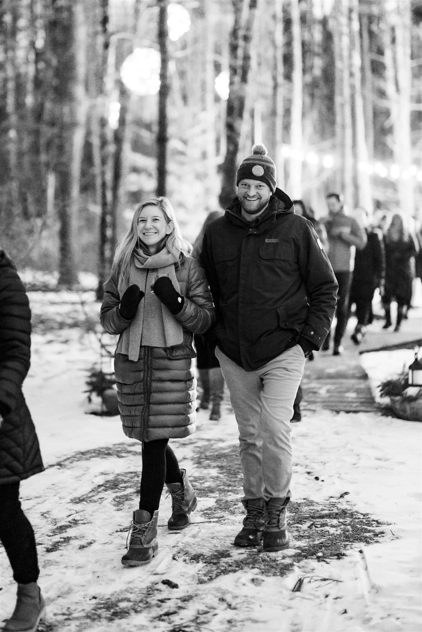 Two smiling people walking on a snowy trail in a forest, wearing winter clothing.