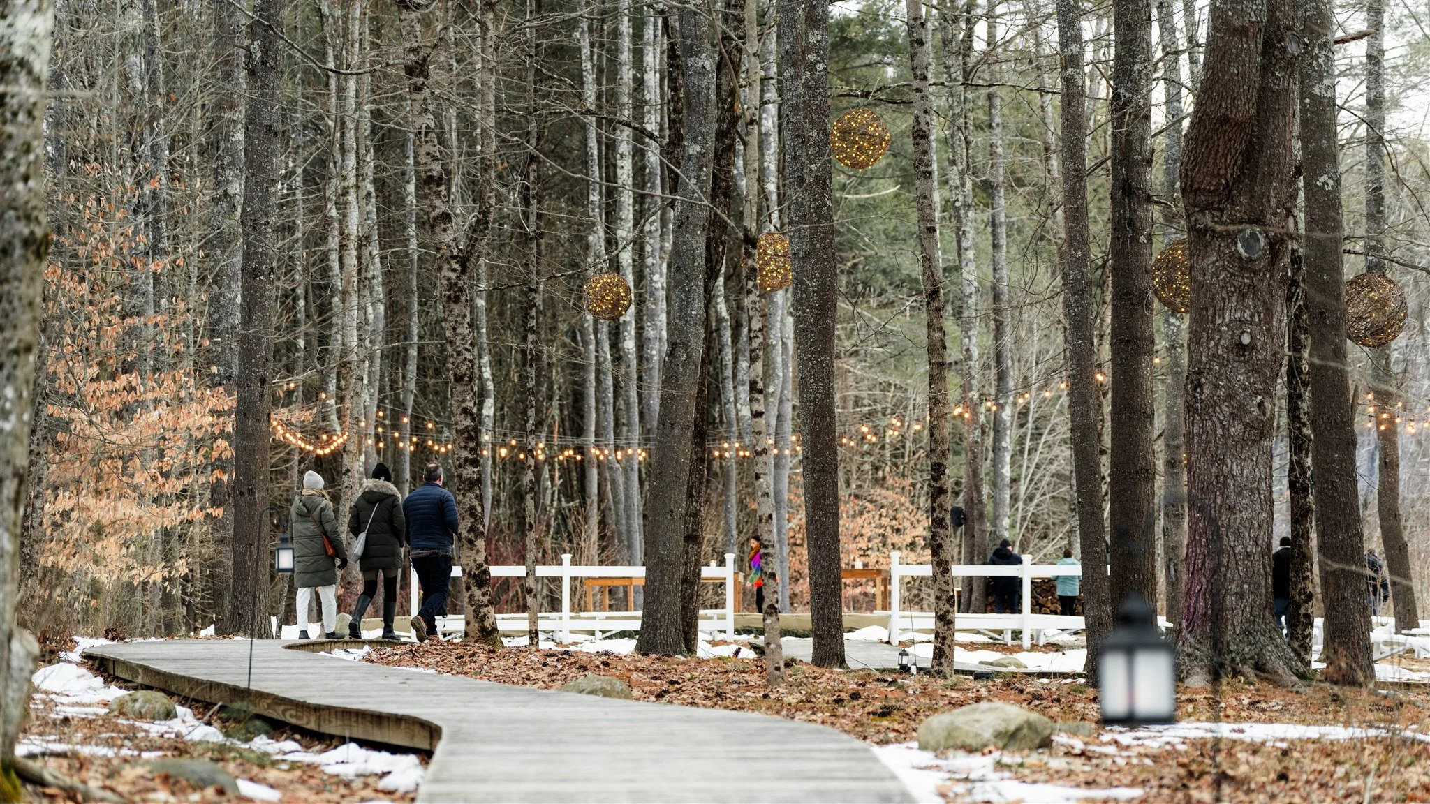 People walking on a wooden path through a forest with string lights and decorative hanging lights, some snow on the ground.