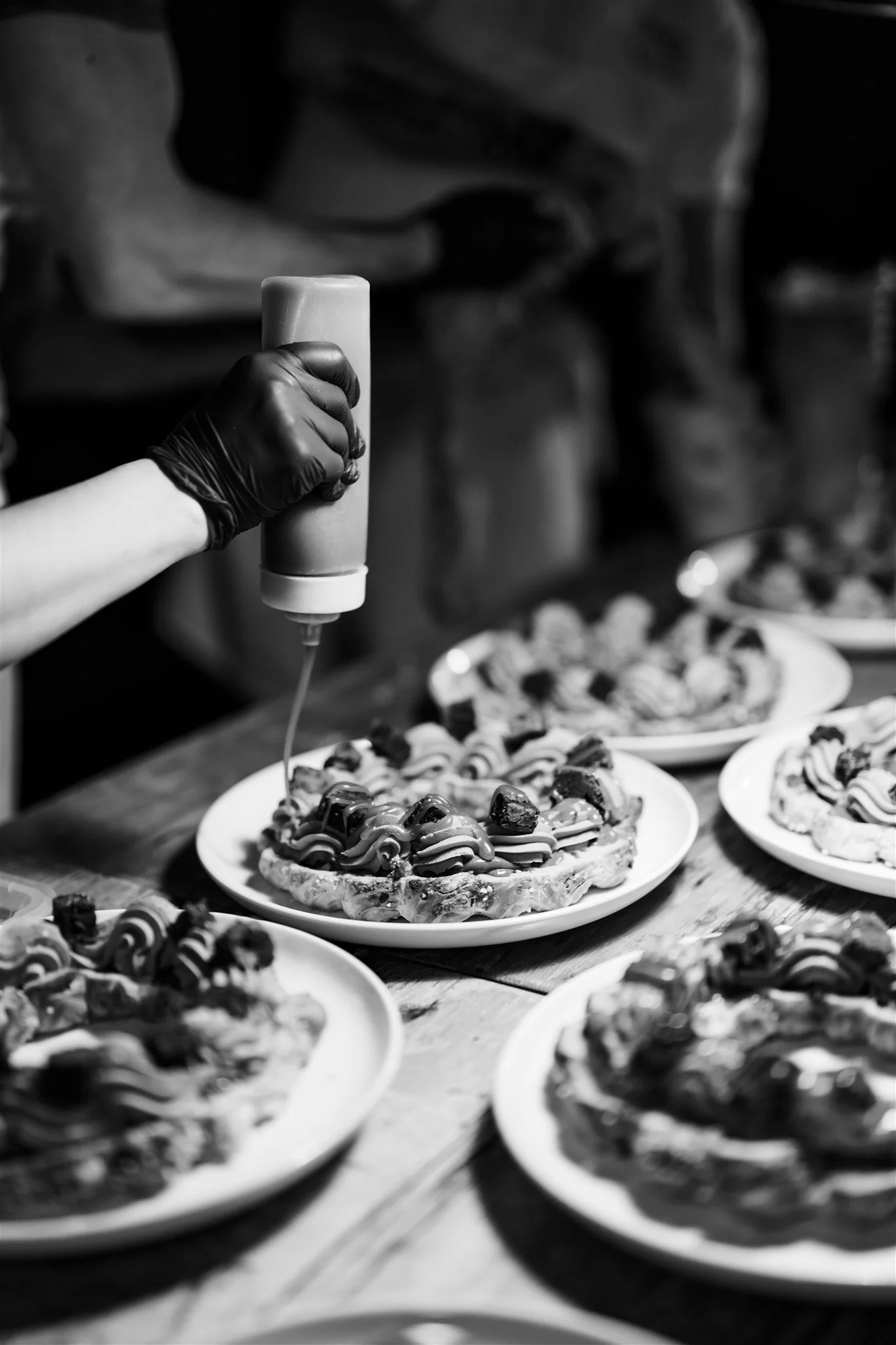 A person wearing black gloves is applying chocolate syrup to multiple plates of small, decorated desserts, with a focus on one plate in the foreground.