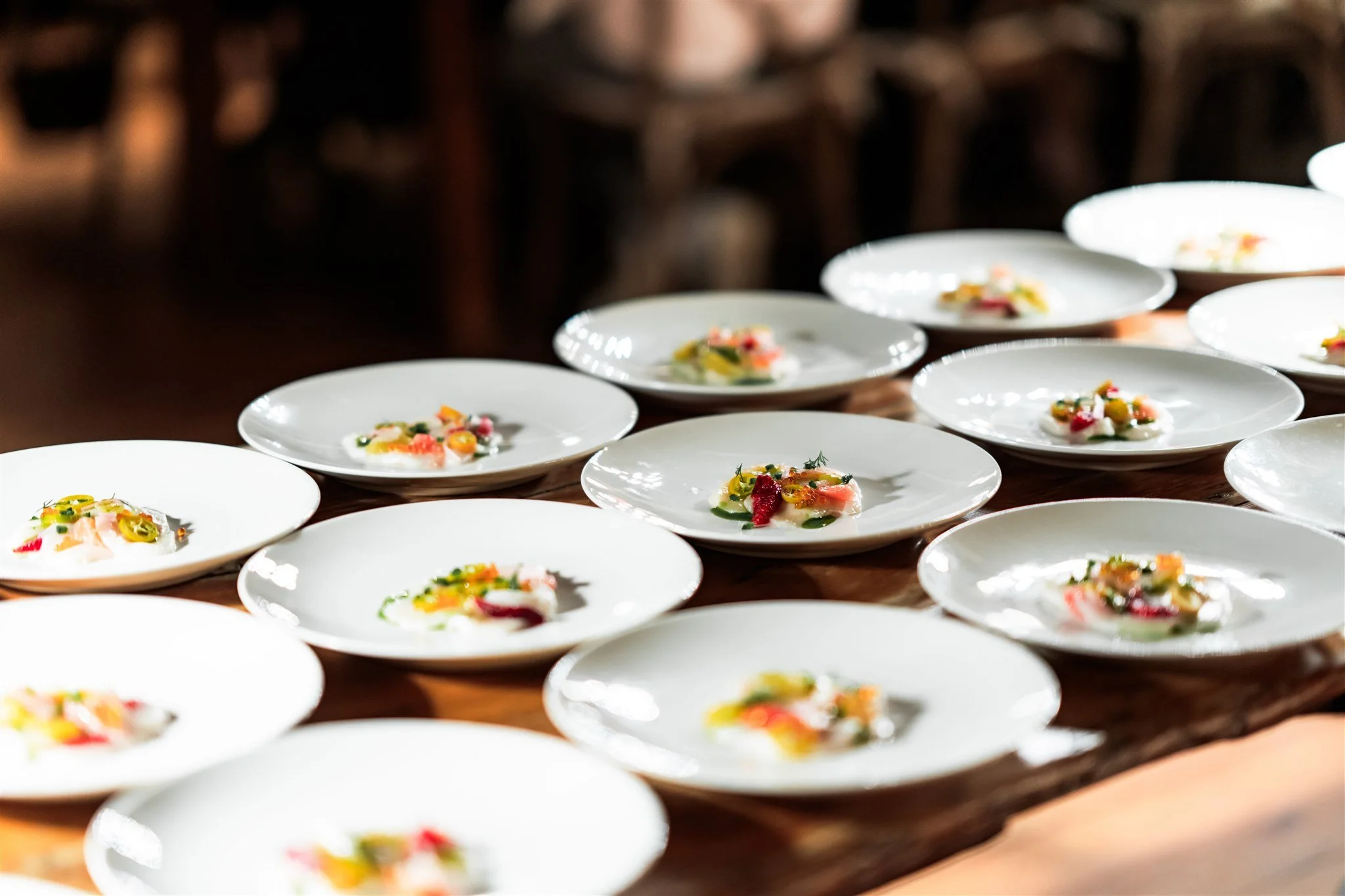 Multiple white plates with small, colorful food appetizers arranged on a wooden table, with chairs blurred in the background.