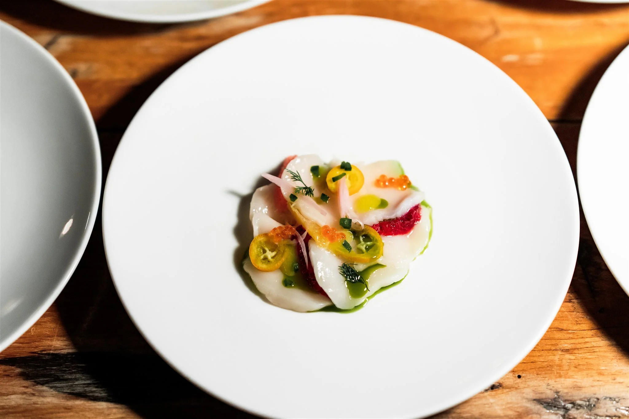 A plated dish with thin slices of raw fish garnished with sliced jalapeños, red onions, yellow fish roe, and fresh herbs on a white plate.
