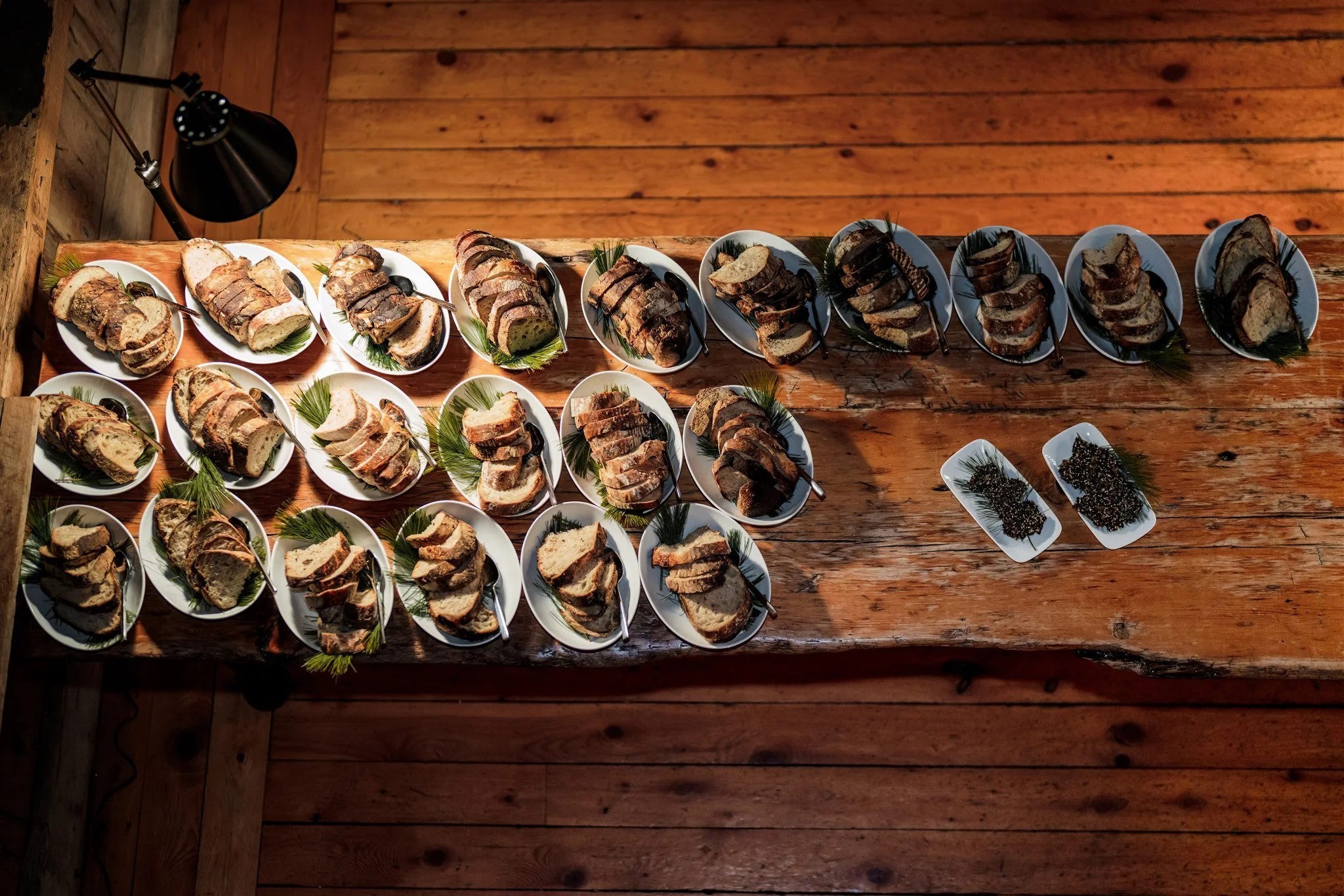 A top-down view of a rustic wooden table with multiple white plates filled with slices of various types of bread and black sesame seeds, decorated with sprigs of greenery, and a black desk lamp positioned on the left side.