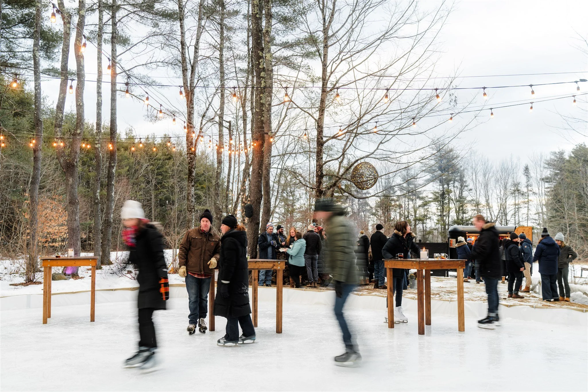 People ice skating on an outdoor rink surrounded by trees with string lights overhead and some winter decor.