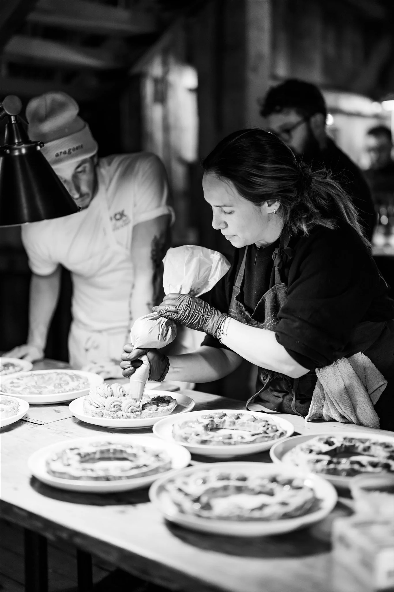 A woman is decorating pizzas on a table with several other people in the background, in a rustic indoor setting.