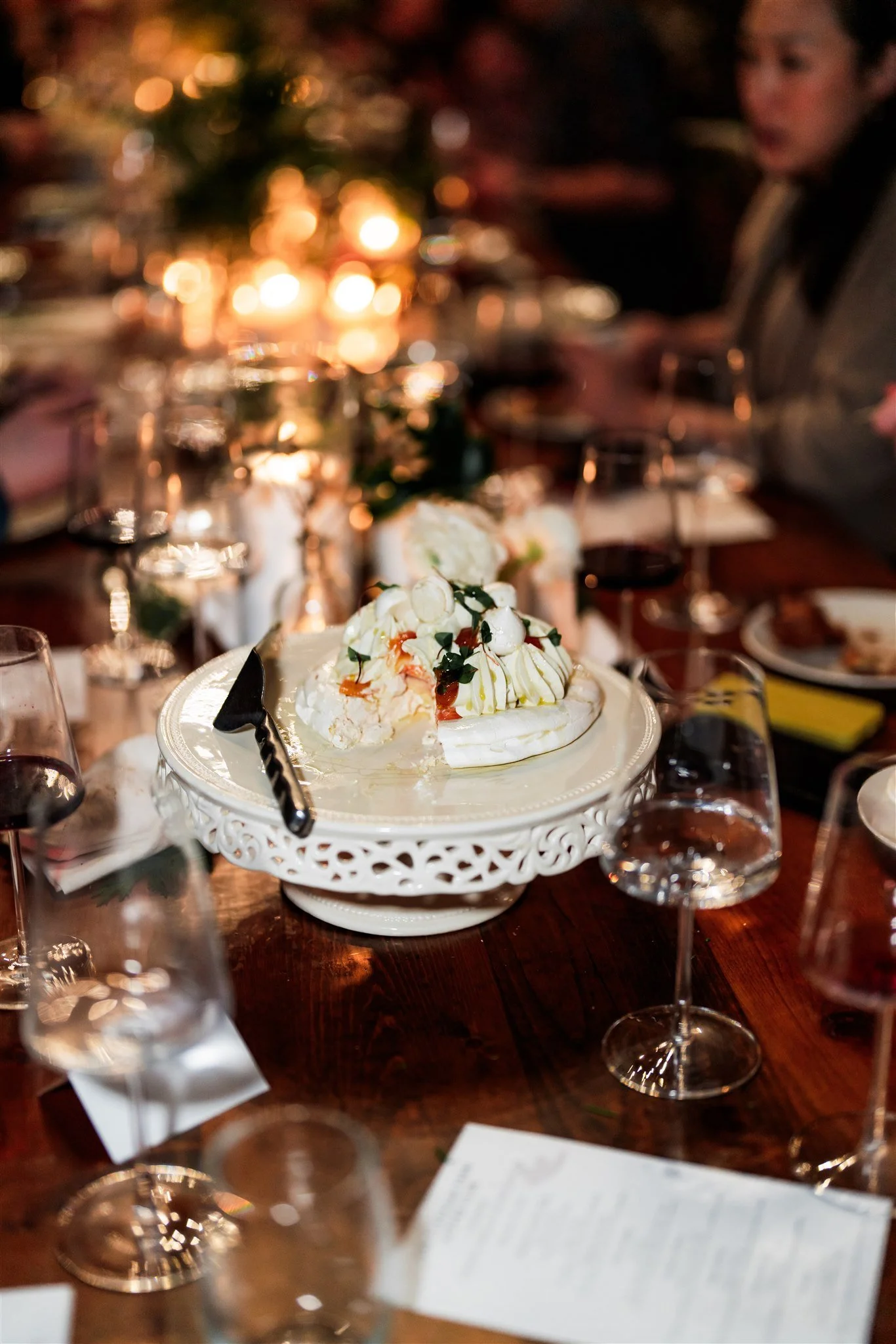 A Christmas dinner table with a partially eaten pavlova cake topped with whipped cream, fruit, and sauce on a white decorative cake stand, surrounded by glasses of red wine, plates, and a Christmas tree in the background.