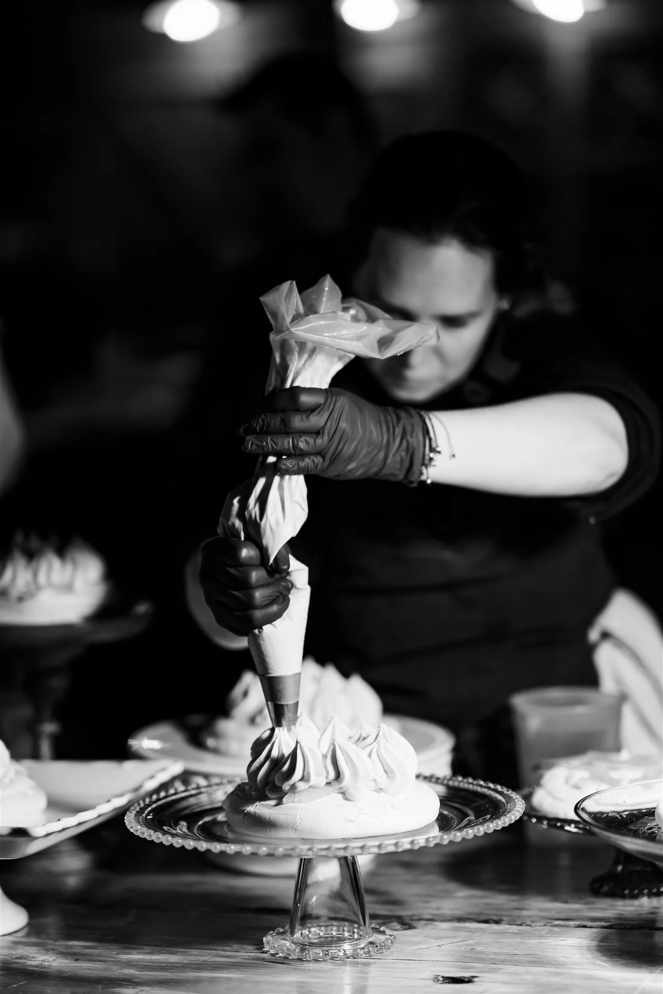 A person wearing black gloves decorating a cake with piped frosting in a piping bag in a bakery or kitchen setting.