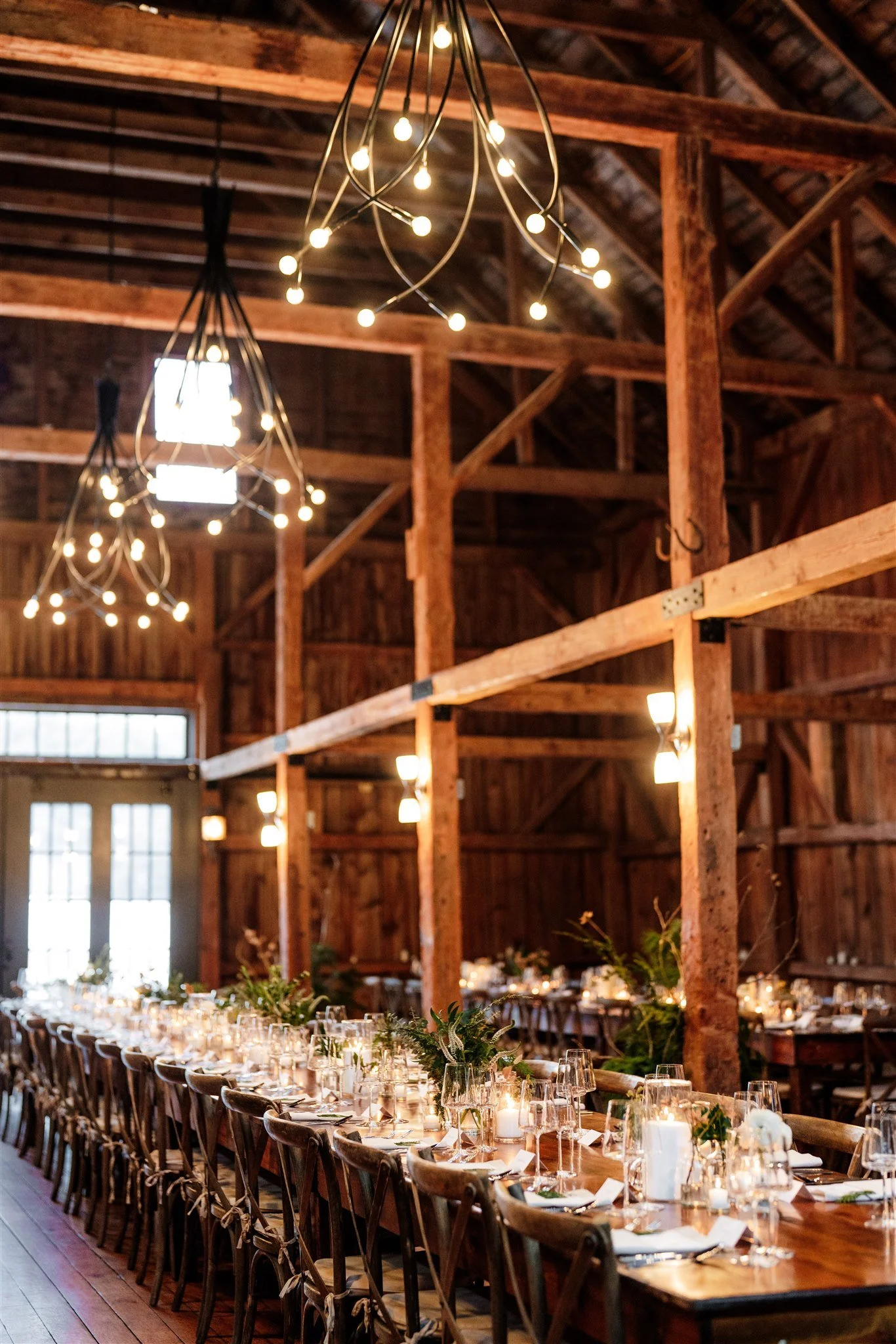 Long banquet table set with glasses, candles, and greenery in a rustic barn with wooden beams and chandeliers.