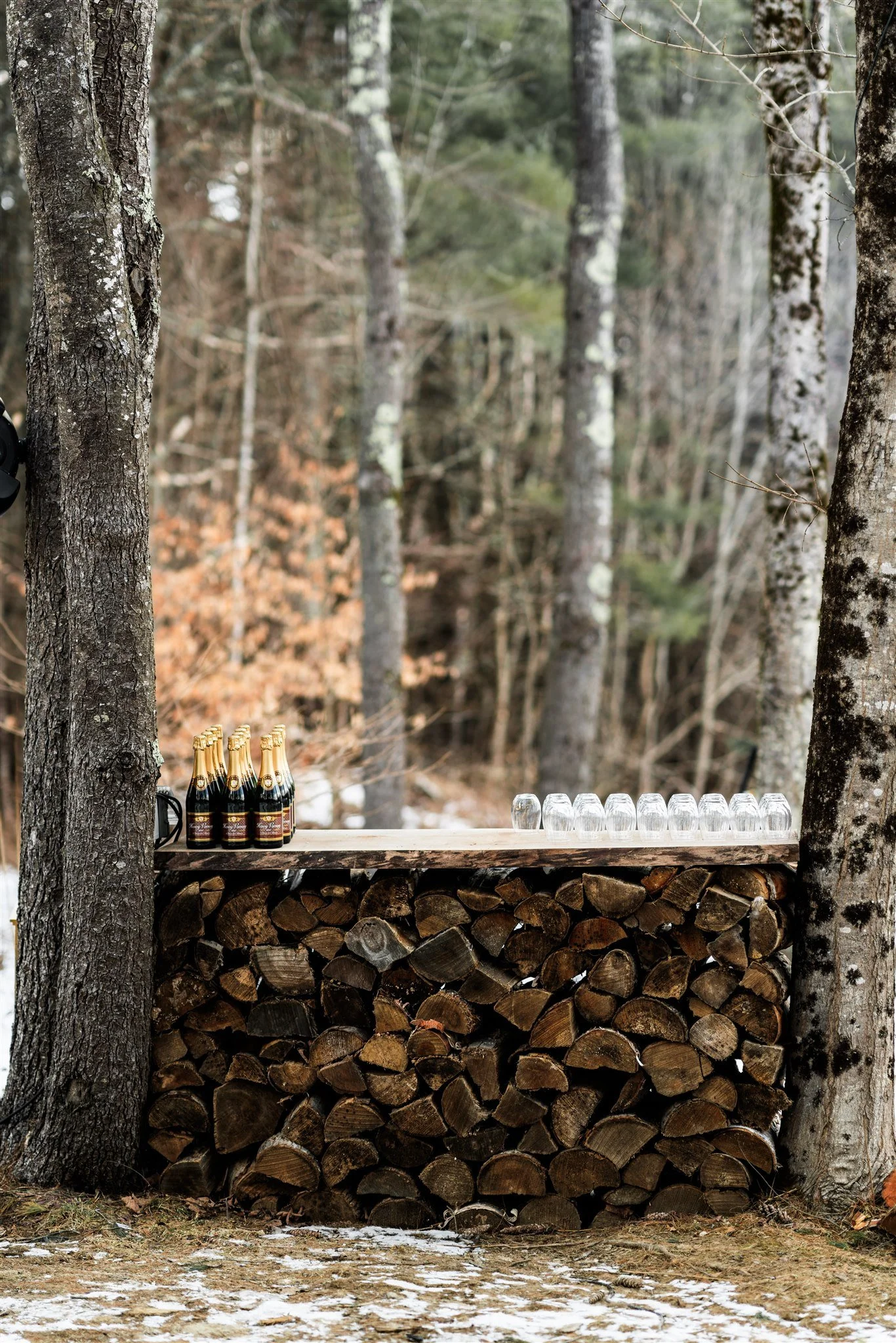 A rustic outdoor bar made from stacked firewood, with bottles of champagne and stacked glasses on top, set in a wooded area with trees.