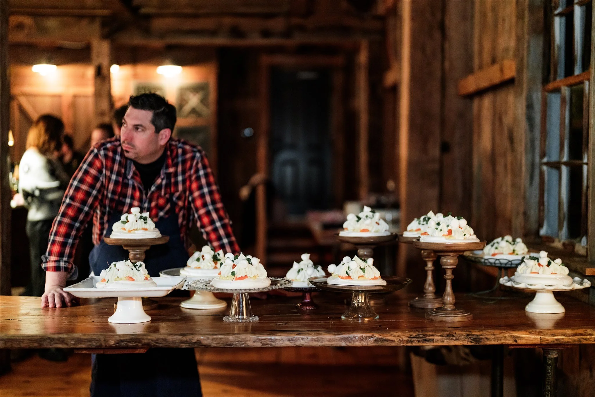 A man in a plaid shirt and apron arranging cakes decorated with white frosting and flowers on wooden and glass cake stands on a rustic wooden table inside a wood-paneled room.