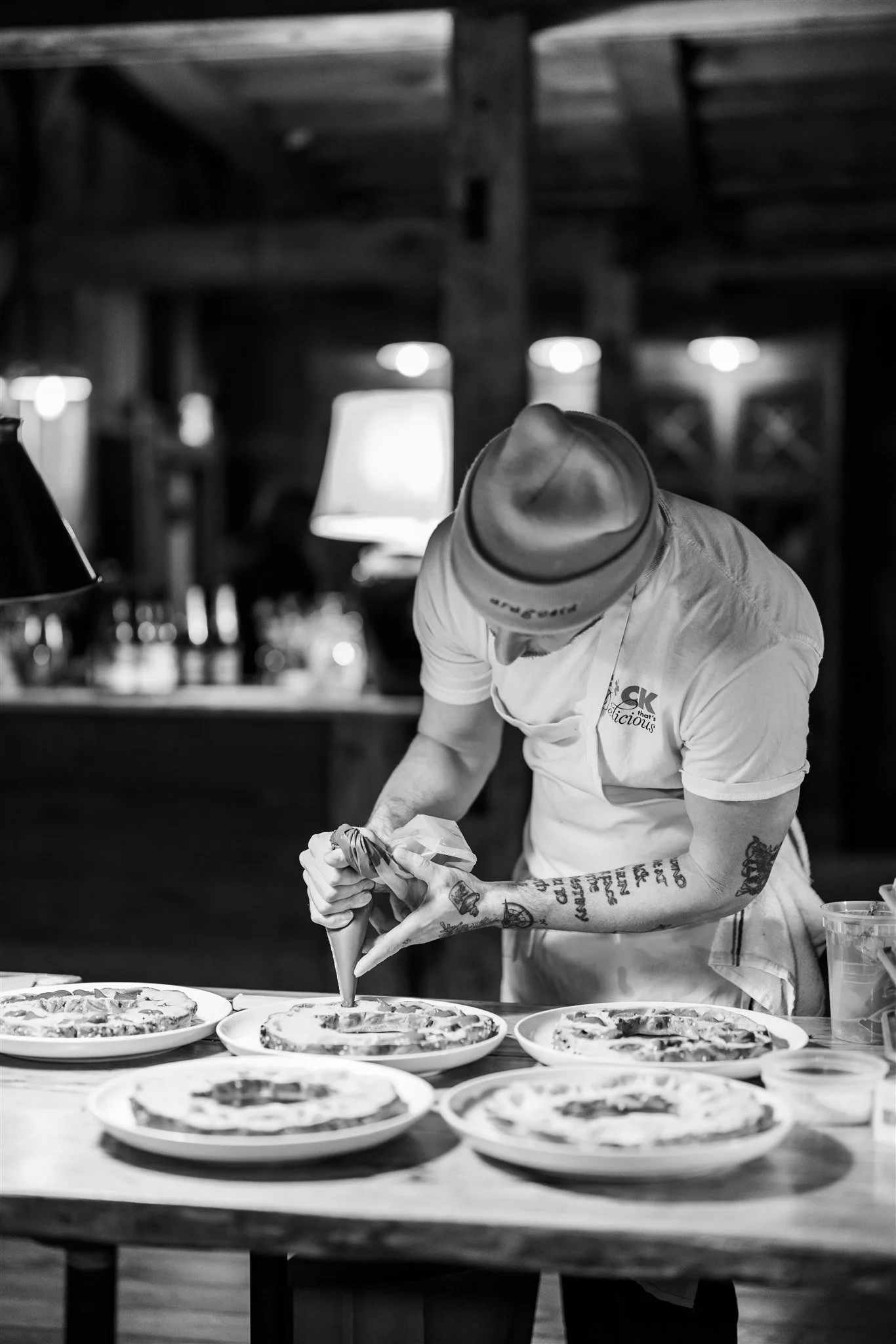 A chef, wearing a hat and white shirt, is preparing multiple pizzas on plates at a rustic restaurant or kitchen. The chef is piping sauce onto a pizza, with several finished pizzas on the table.