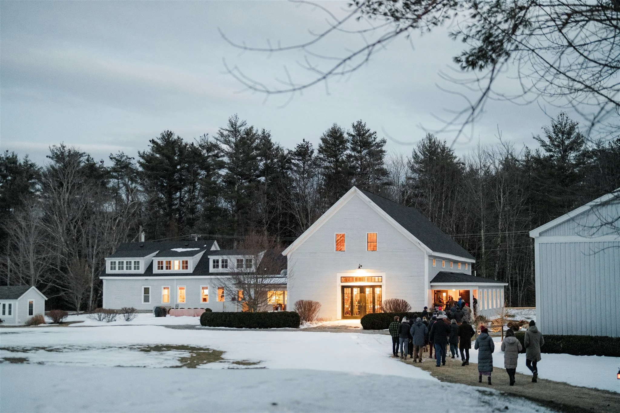 Groups of people walking toward a large white building in a winter setting, with snow on the ground and trees in the background, warm lights glowing from inside.