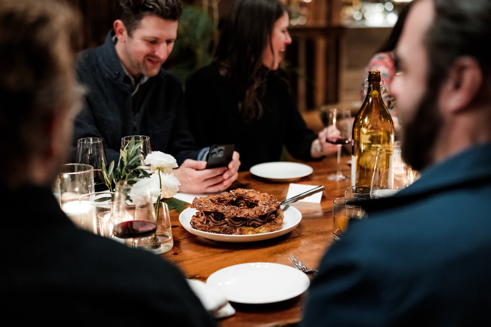People seated around a dinner table with a chocolate dessert, wine glasses, and a large brown bottle, in a cozy, wood-paneled setting.
