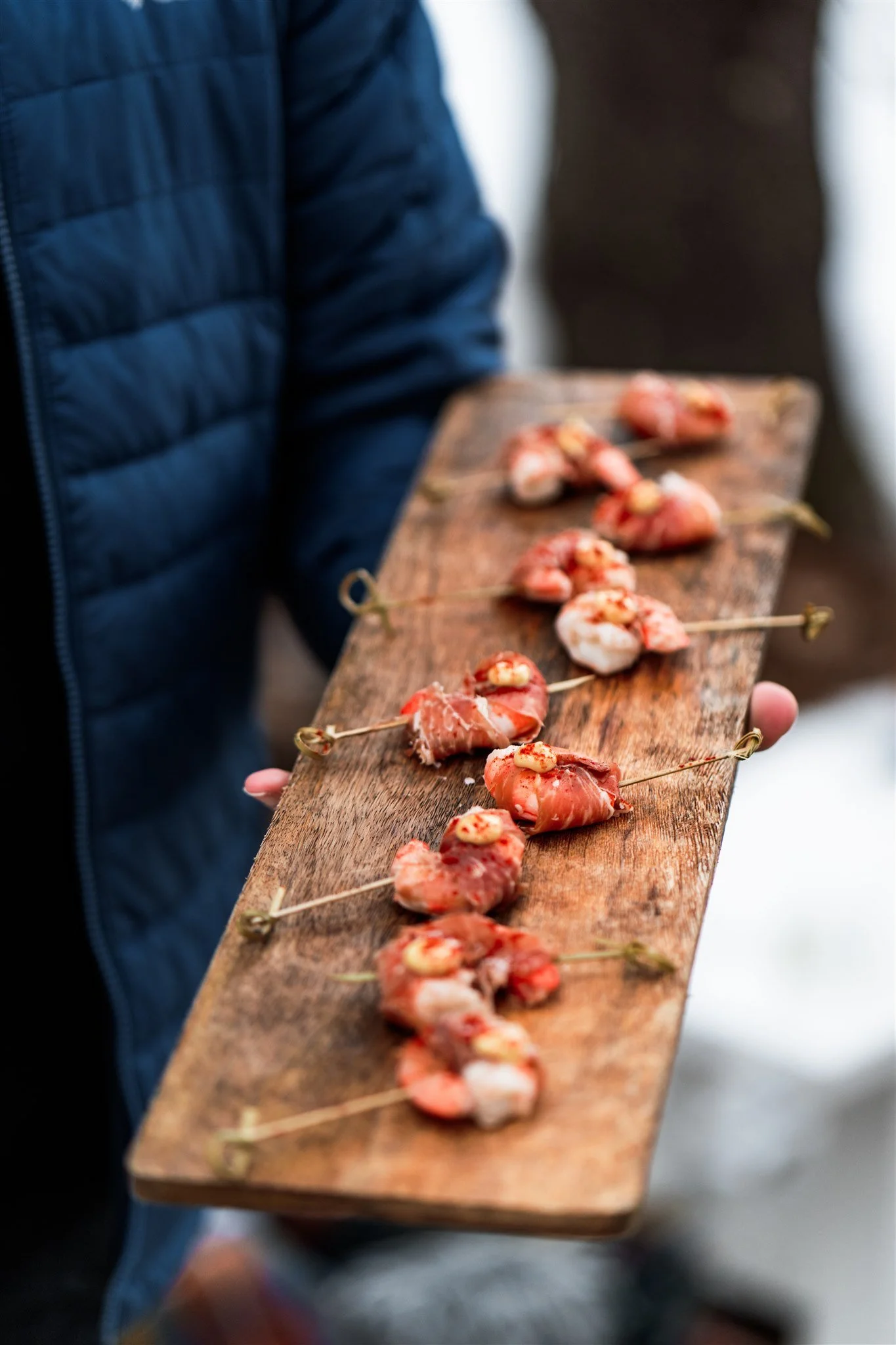Person holding a wooden serving plank with shrimp wrapped in prosciutto skewered on small wooden sticks.