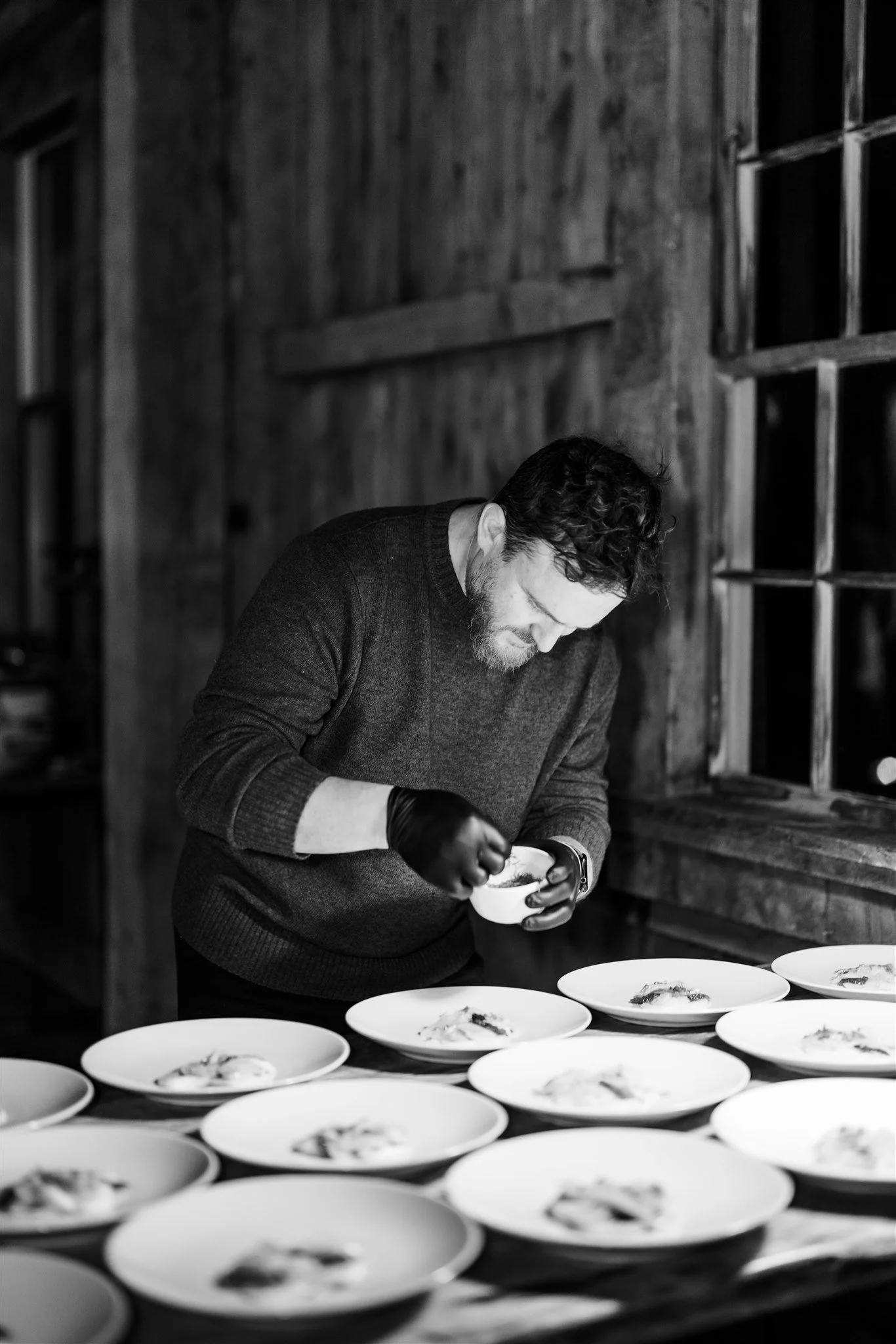 A man with curly hair, wearing a dark sweater and black gloves, is arranging or garnishing dishes on a table in a rustic, wood-paneled room.