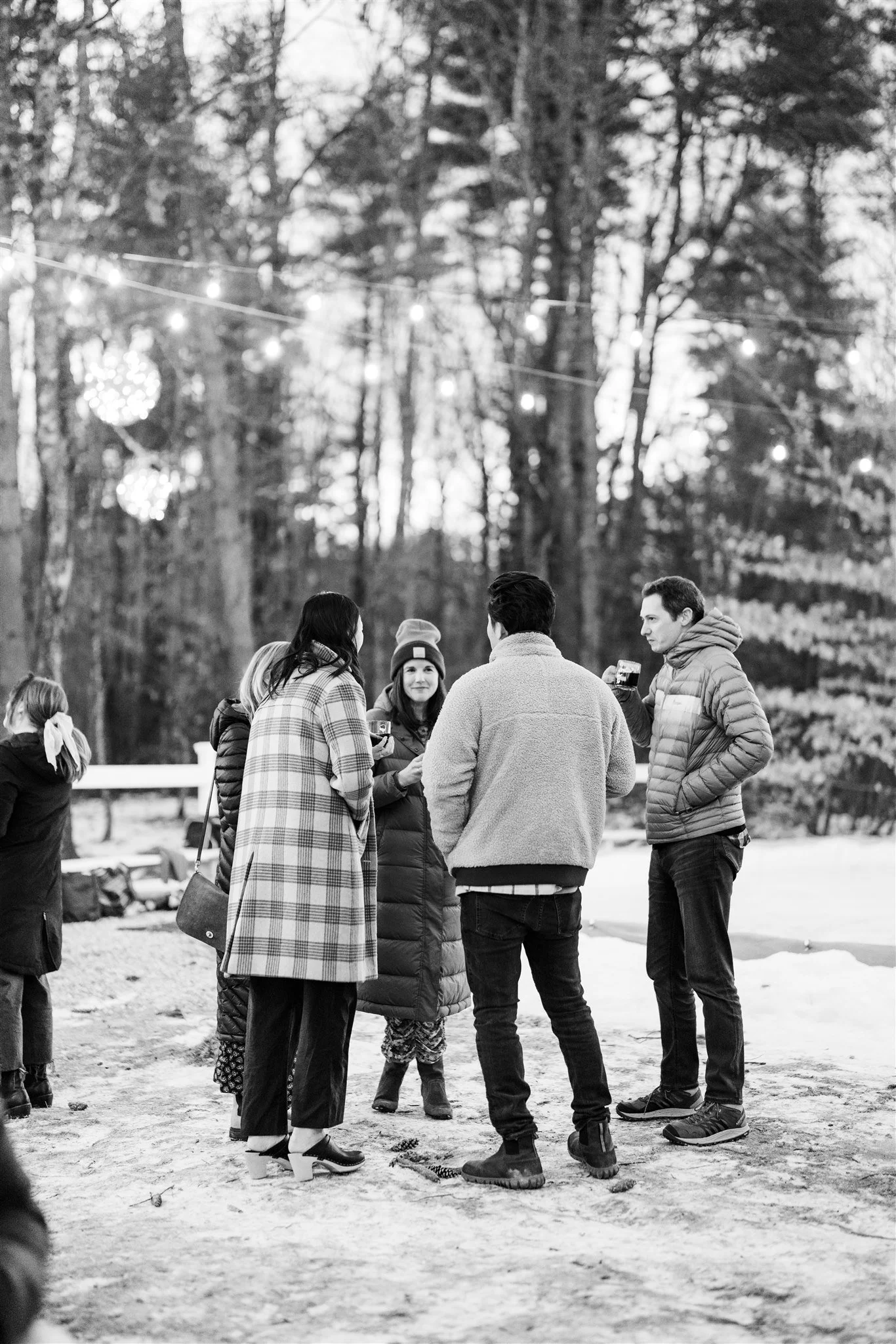 People standing outdoors in winter, talking and holding drinks, with string lights overhead and trees in the background.