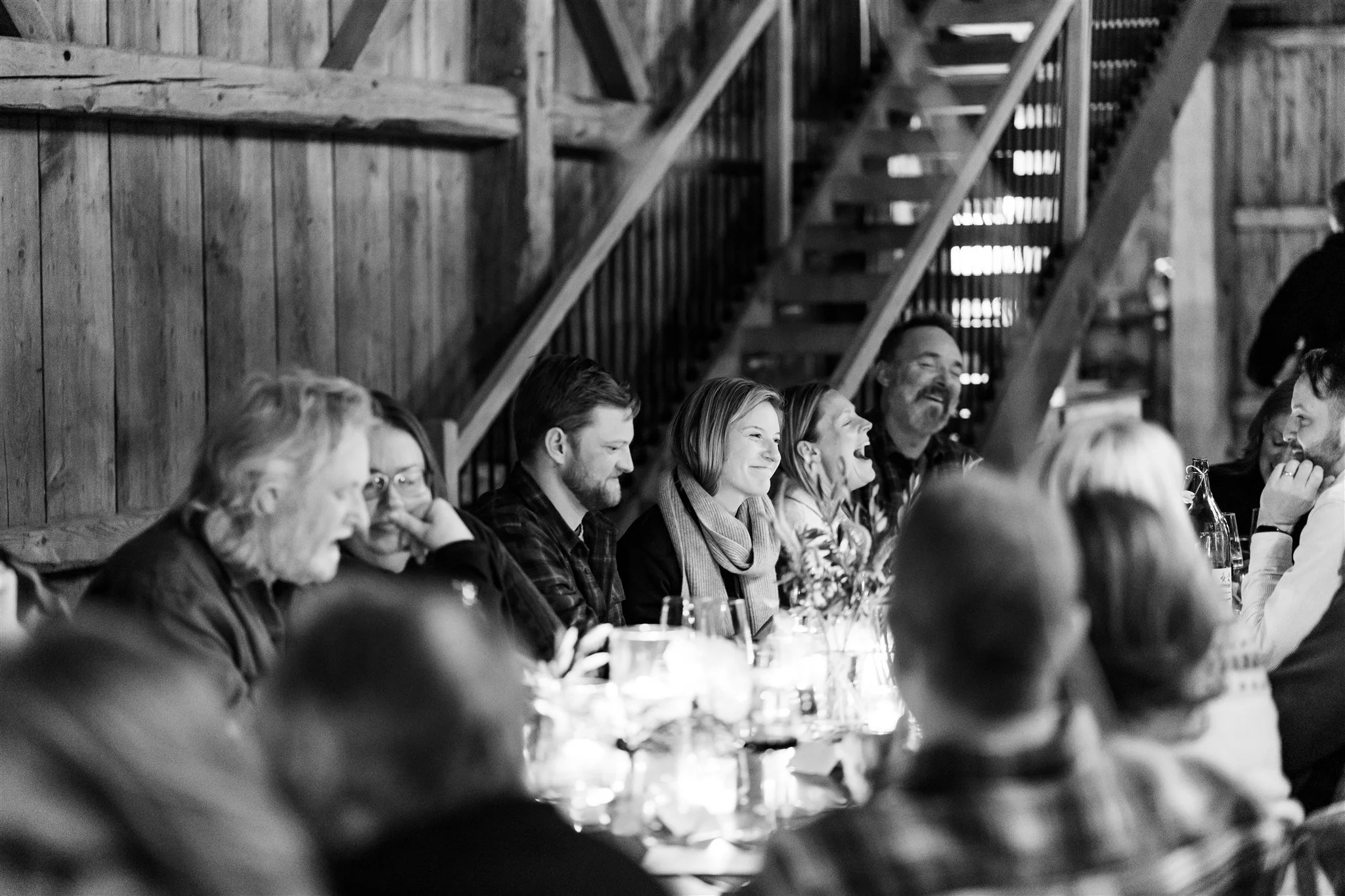 People sitting at a long table in a rustic indoor setting, enjoying a meal and laughing, with wooden walls and stairs in the background.