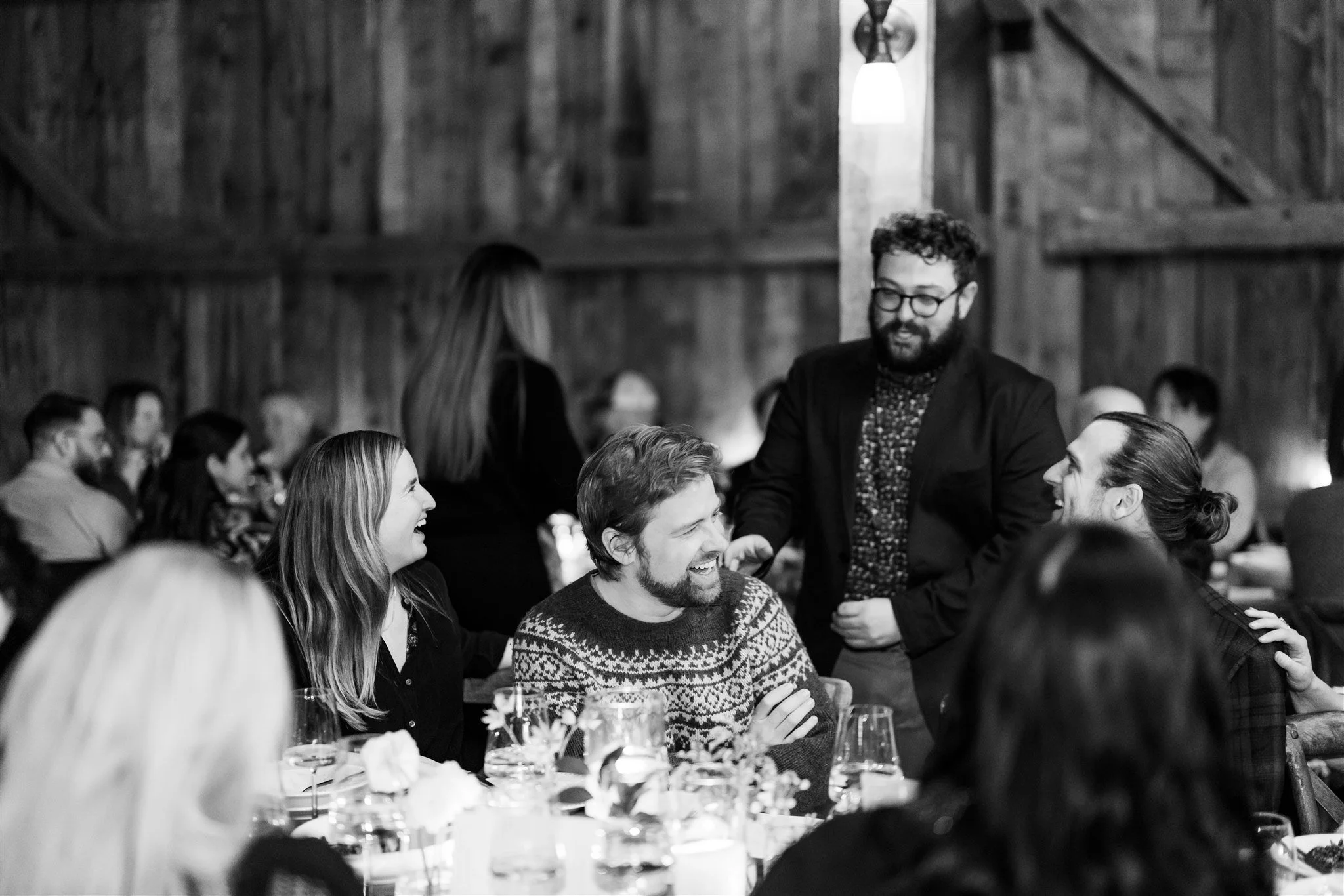 A group of people at a social gathering, sitting around a table, smiling and laughing, with one man standing and engaging in conversation in a wooden interior setting.