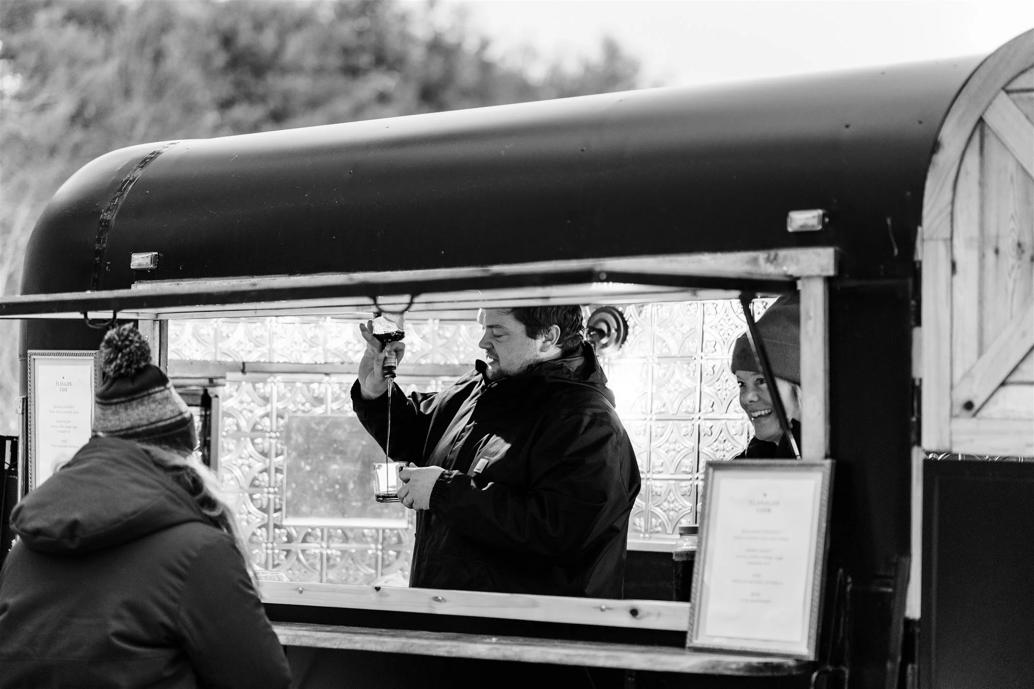 People at a food truck, with one man pouring a drink and another woman smiling inside.