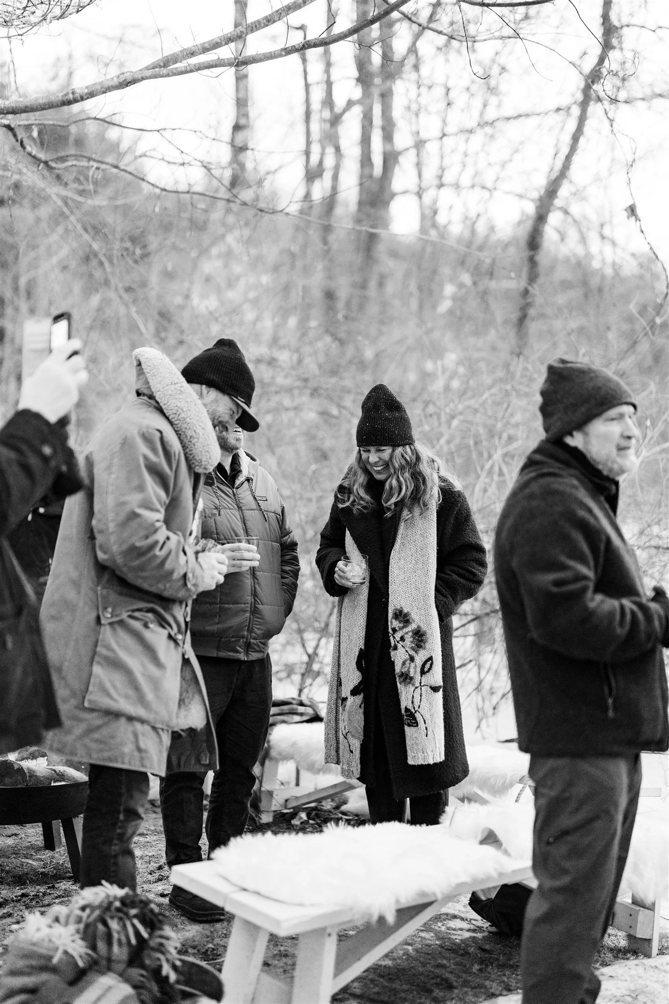 A group of five people standing outdoors in winter, wearing warm clothing and beanies, engaging in conversation and taking photos in a wooded area.