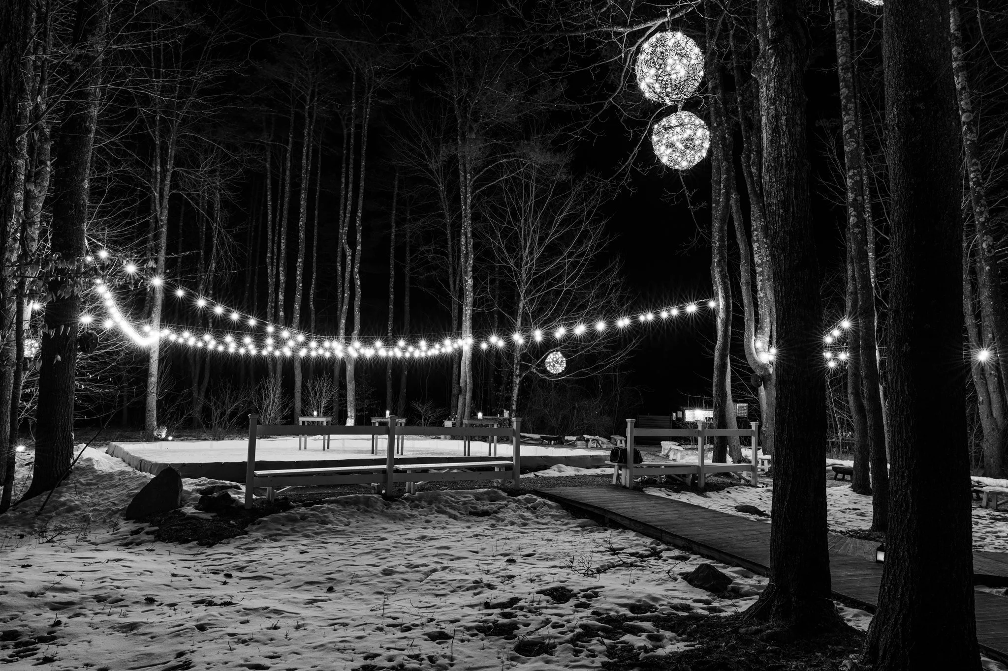 Night scene in a snow-covered forest with string lights hanging between trees, illuminated spherical lanterns hanging from above, and a small wooden platform or stage surrounded by trees.