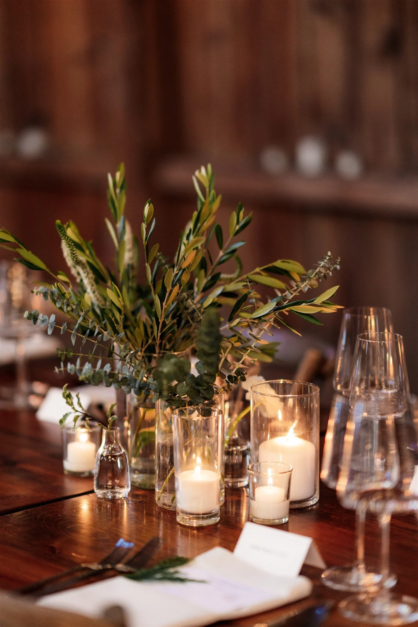A table decorated with a centerpiece of greenery, surrounded by candles in glass holders, with empty wine glasses and table settings.