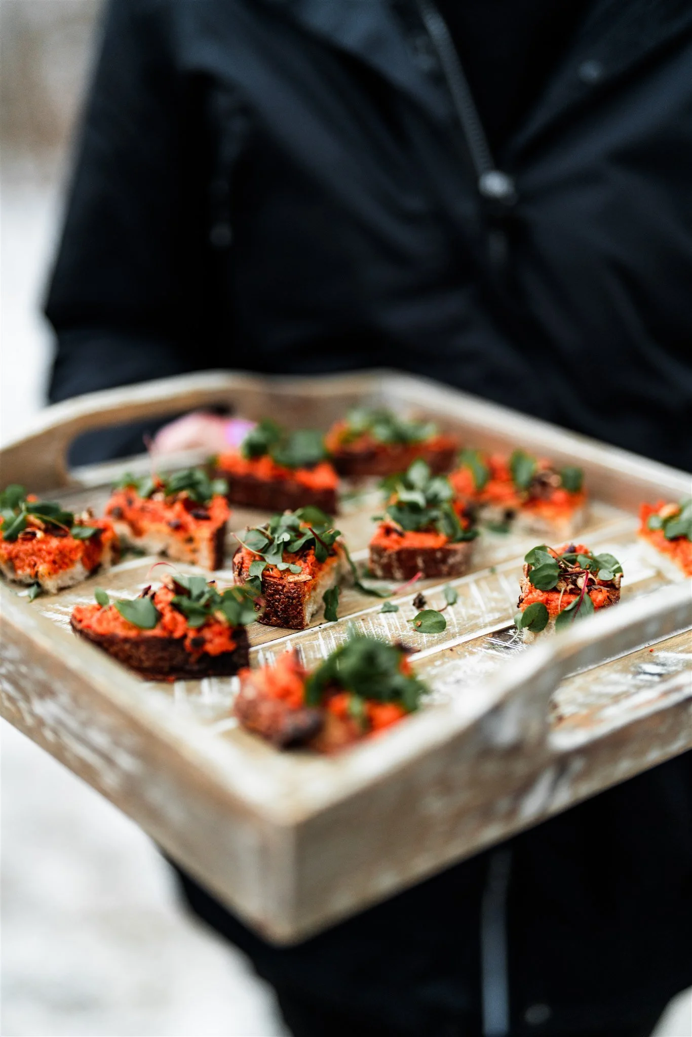 Person holding a tray of bite-sized appetizers topped with greens, served on a silver tray in an outdoor setting.