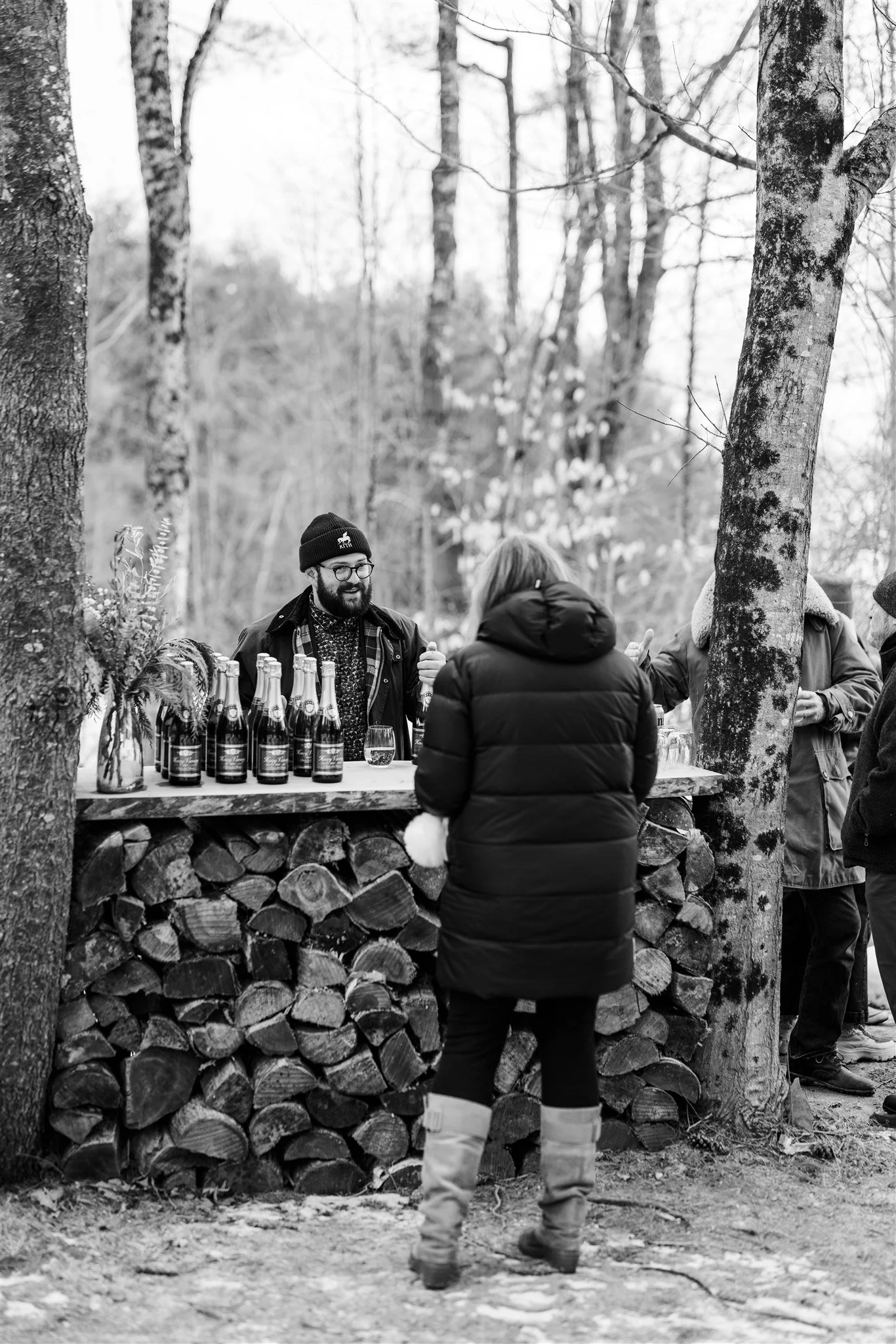 People at an outdoor bar with bottles of beverages, surrounded by trees in a forest setting, in black and white.