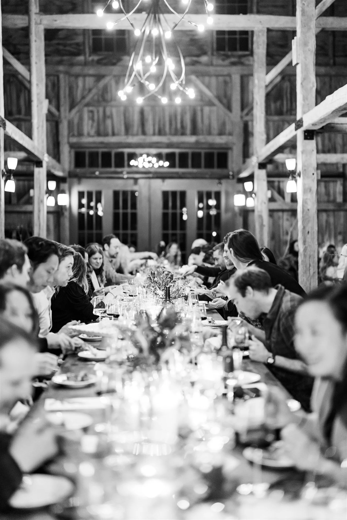 A black and white photo of a long dinner table with people dining in a rustic wooden barn, illuminated by chandeliers and wall-mounted lights.