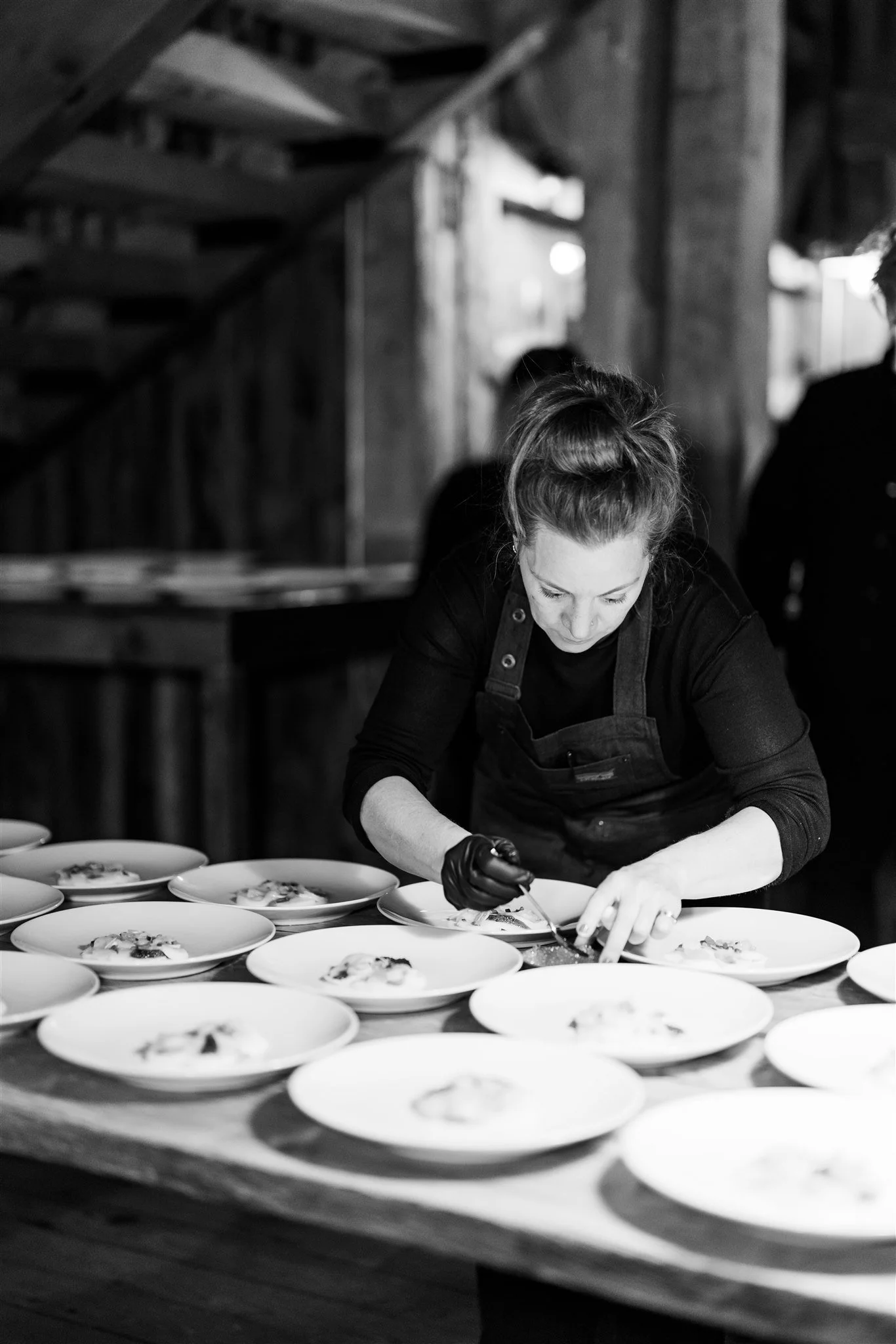 A woman wearing a black long-sleeve shirt, a black apron, and a glove on her right hand, leaning over a table and carefully plating dishes in a rustic setting. The table is lined with multiple plates with food, and the background features wooden wall