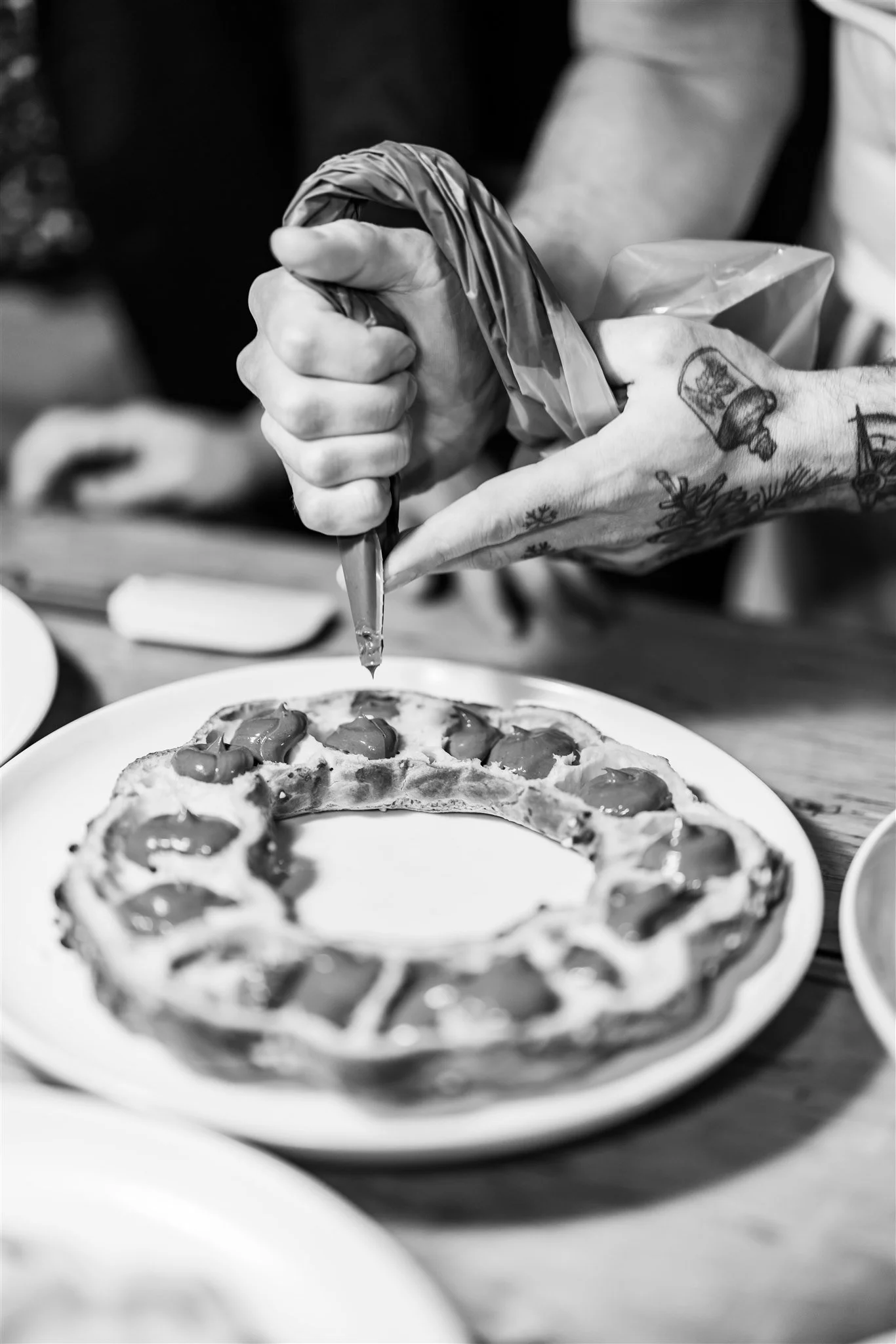 A tattooed hand holds a piping bag and decorates a pizza with toppings on a white plate.