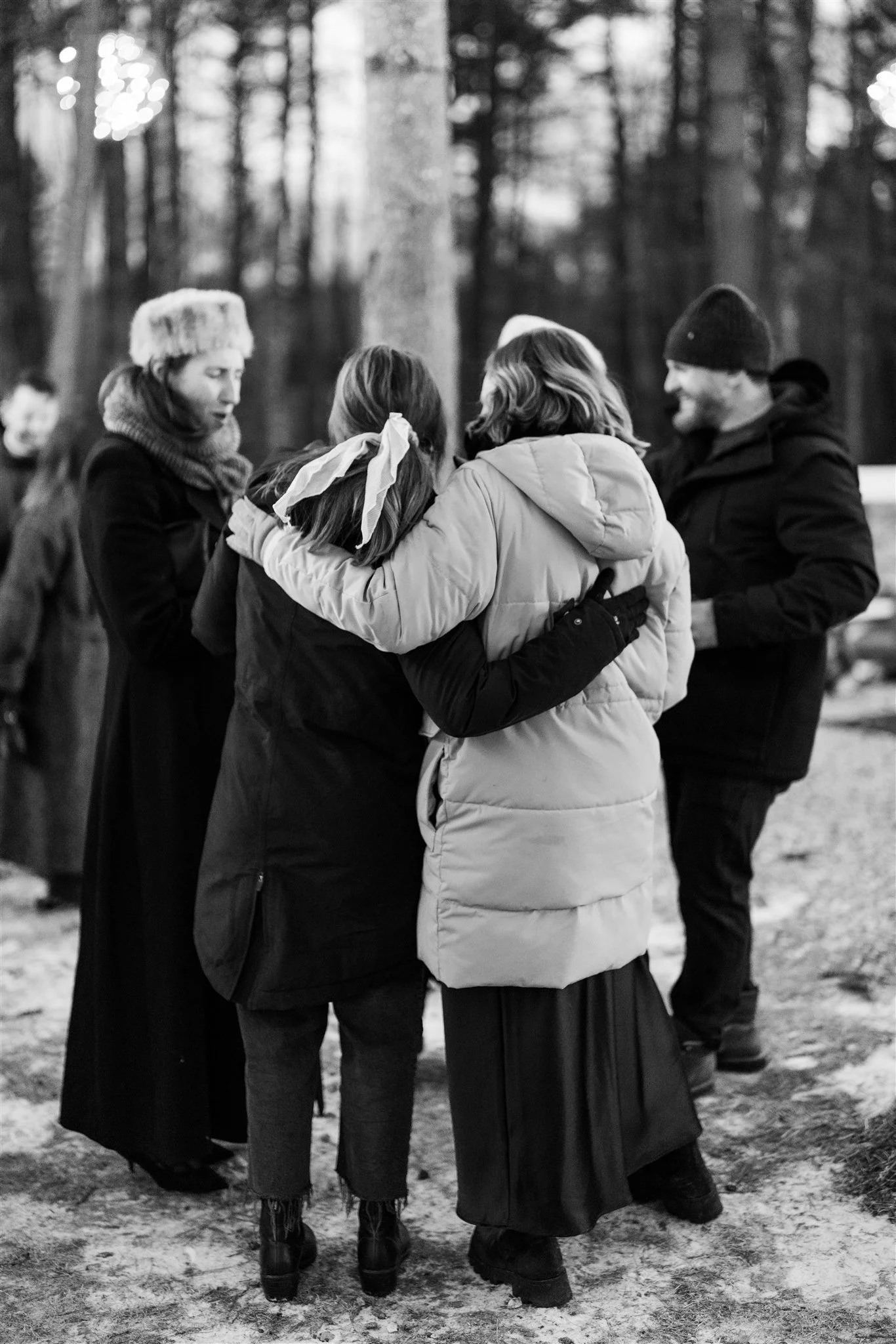 A group of people, mostly women, embracing and comforting each other outdoors in a wooded area during winter, with some wearing warm jackets and hats.