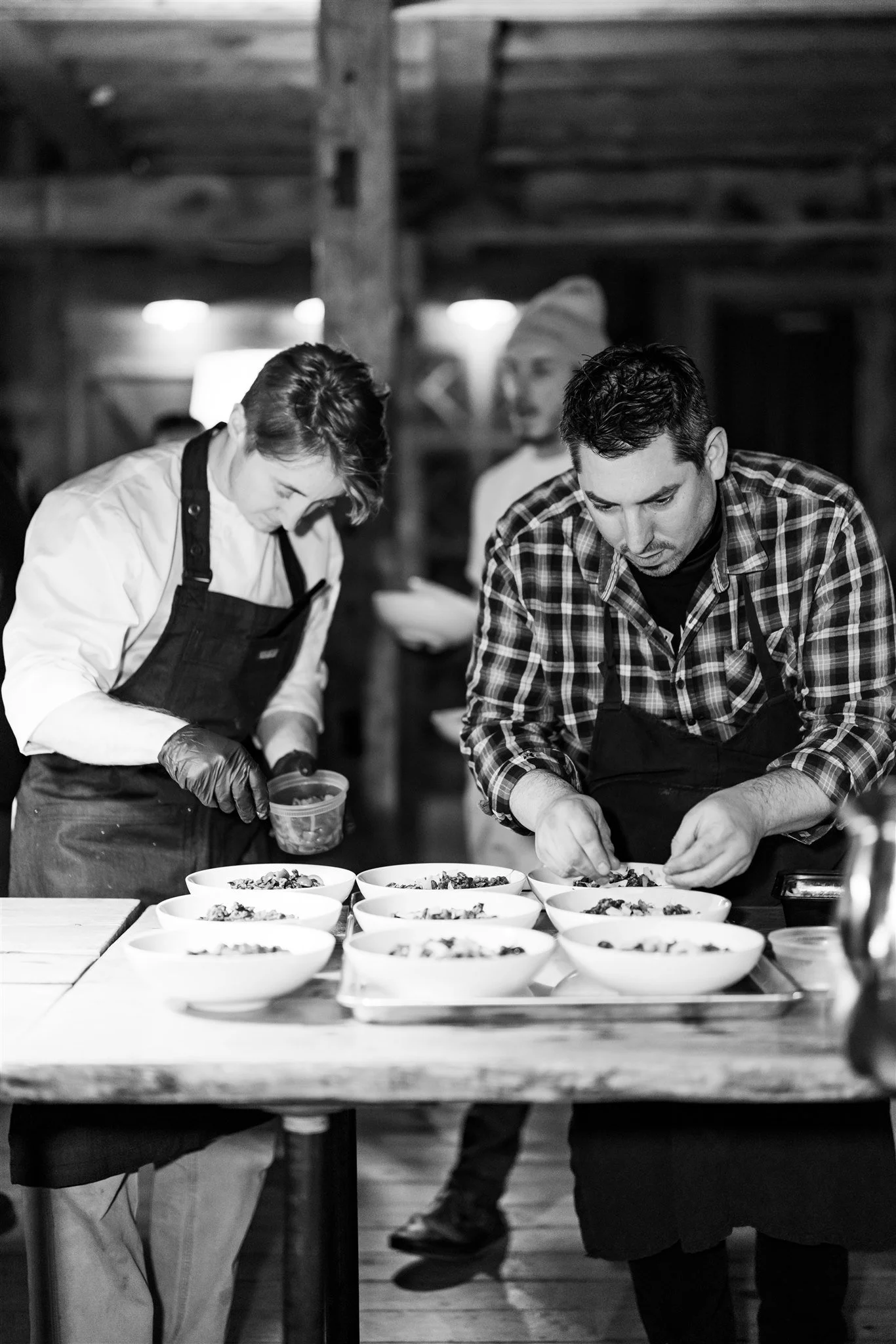 Two chefs, a woman and a man, preparing dishes in a rustic kitchen with bowls of food on the table.