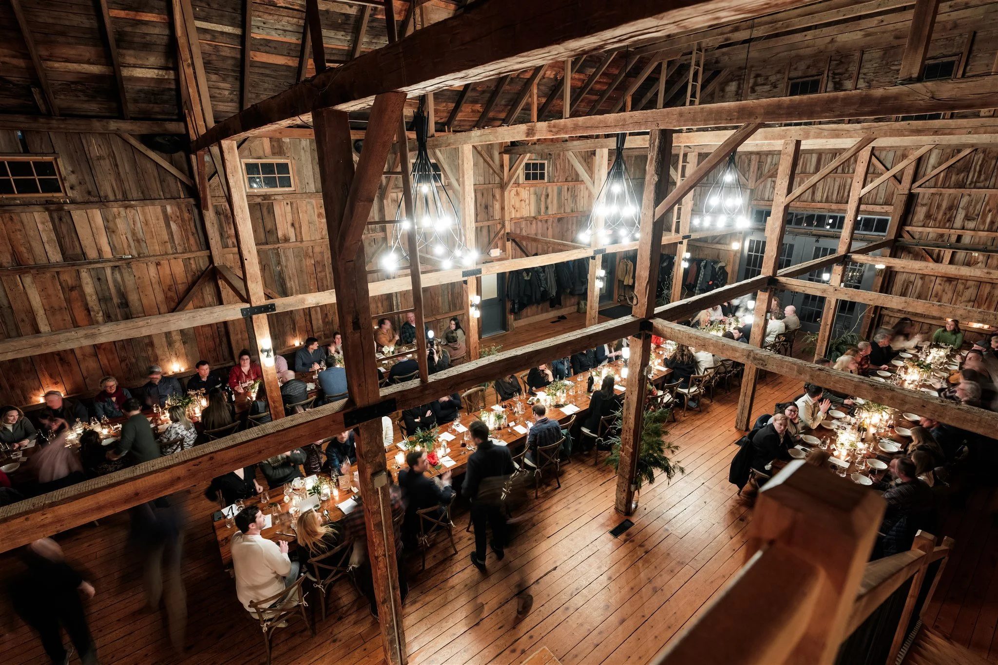 A large rustic barn-style wedding reception with guests seated at long tables, wooden beams, chandeliers, and warm lighting.