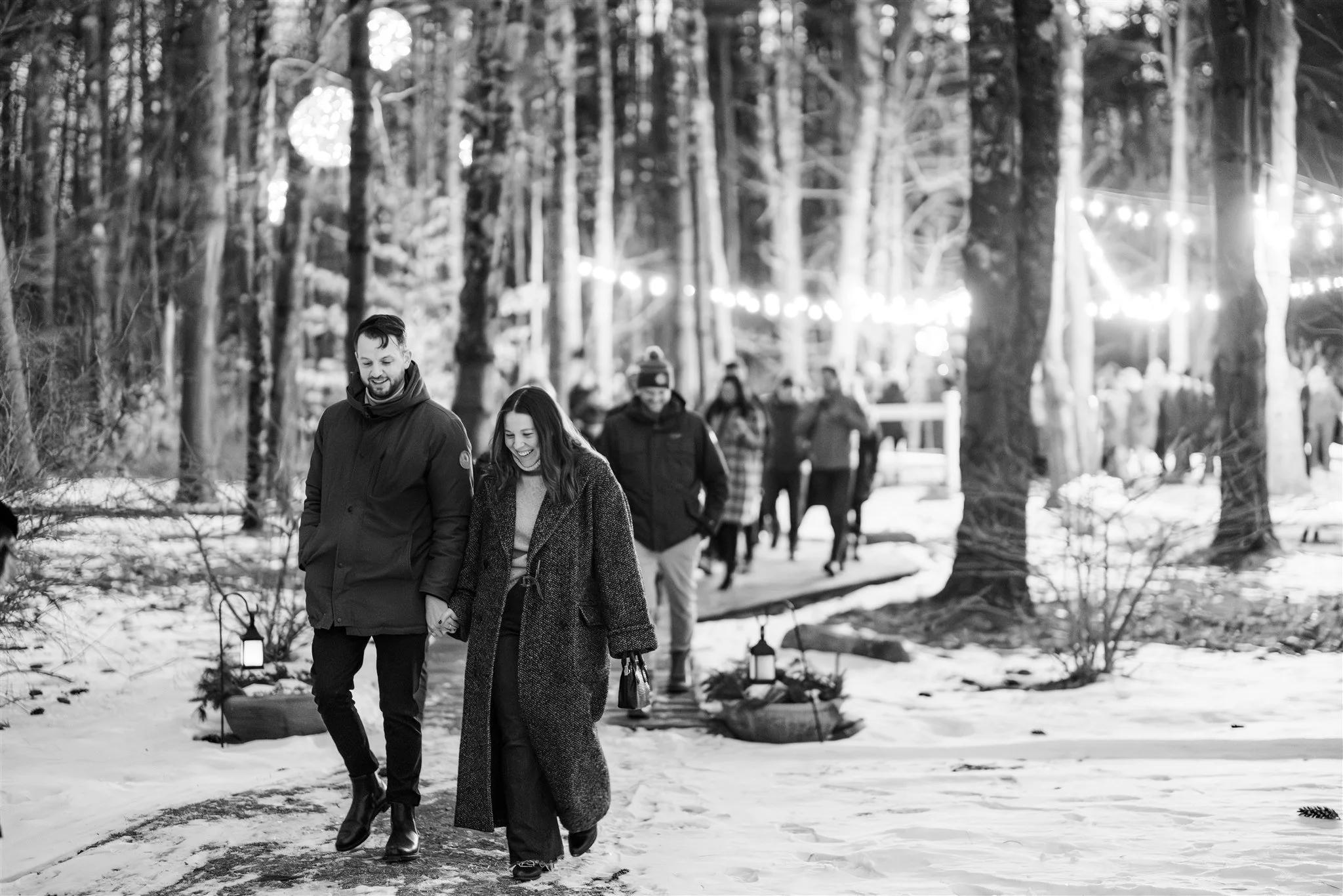 People walking through a snowy forest at night with string lights hanging among the trees, some smiling and holding hands.