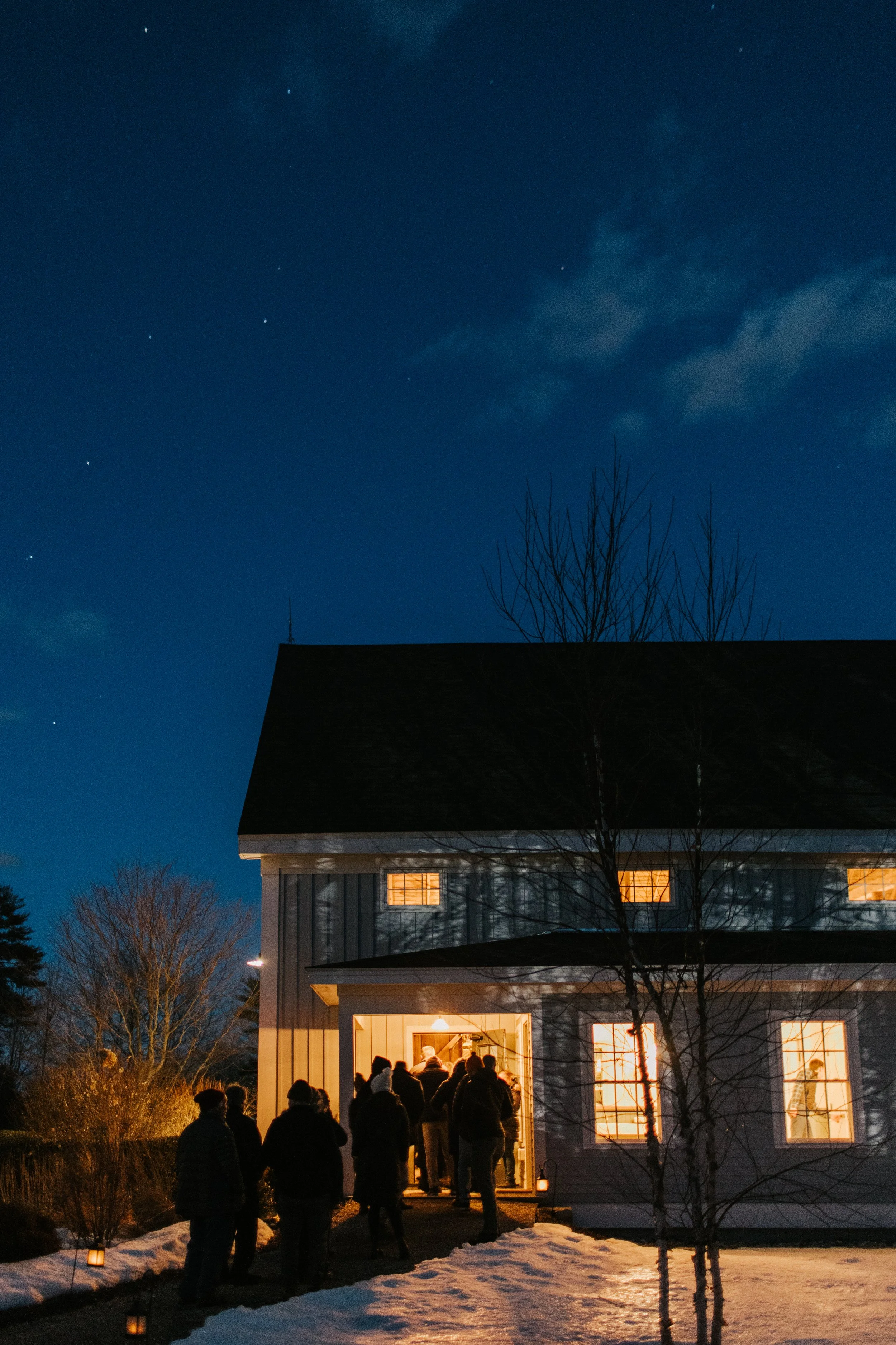 People gathered outside a house at night, warmly lit, with a starry sky overhead and snow on the ground.