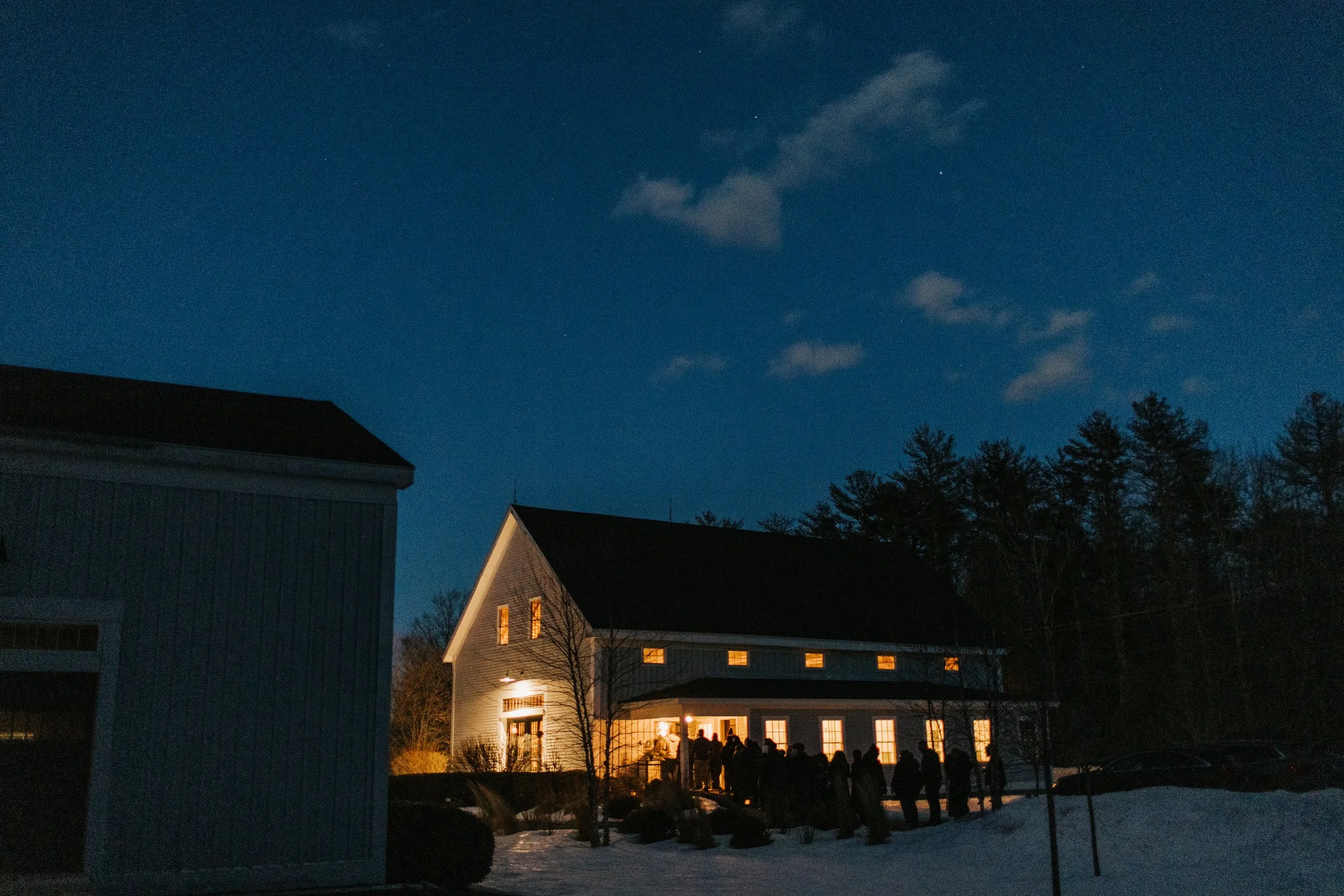 People gathering outside a well-lit house at night with snow on the ground and a dark sky overhead.