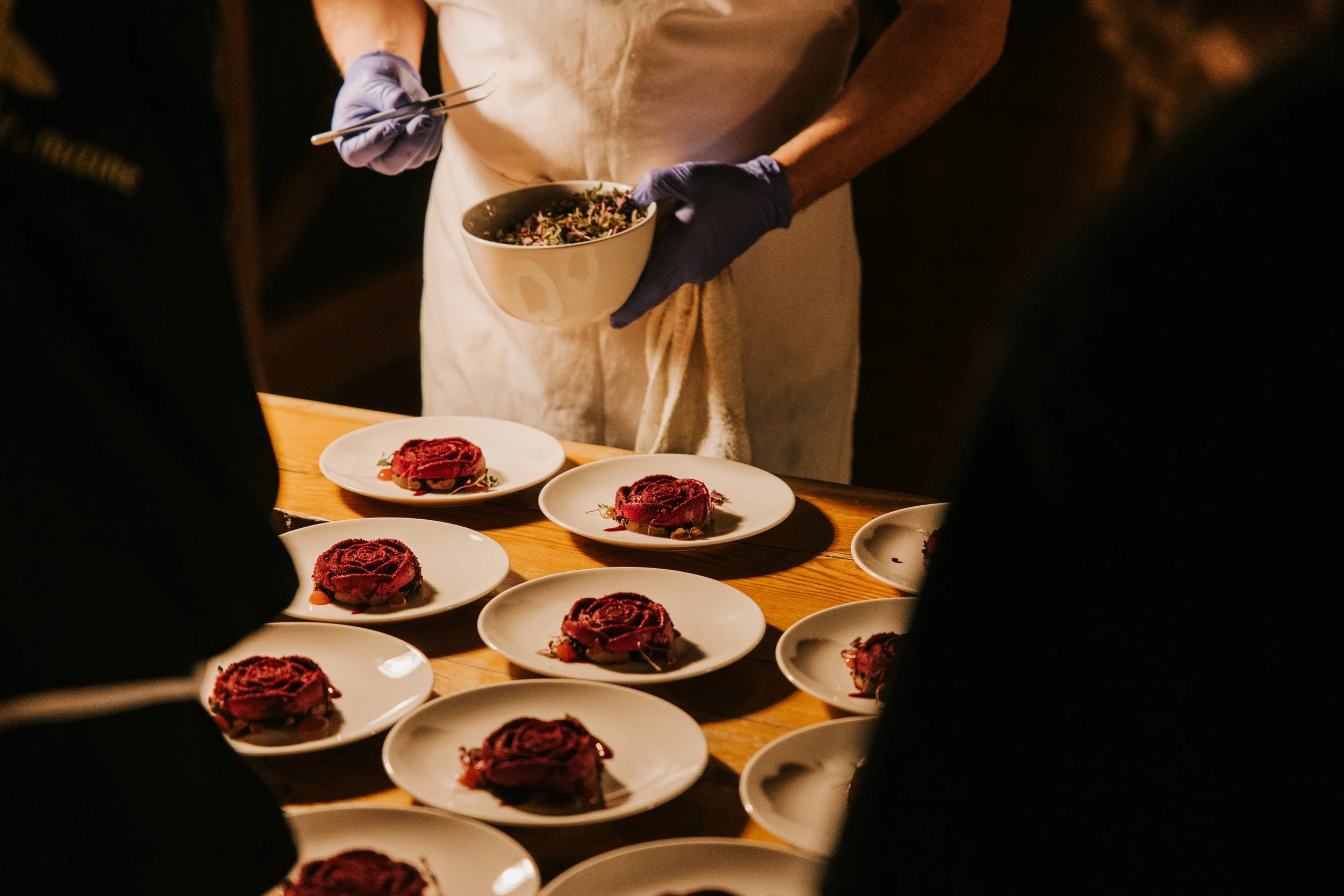 A chef plating multiple small desserts on white plates while holding a bowl of salad. The scene appears to be in a professional kitchen or intimate dining setting.