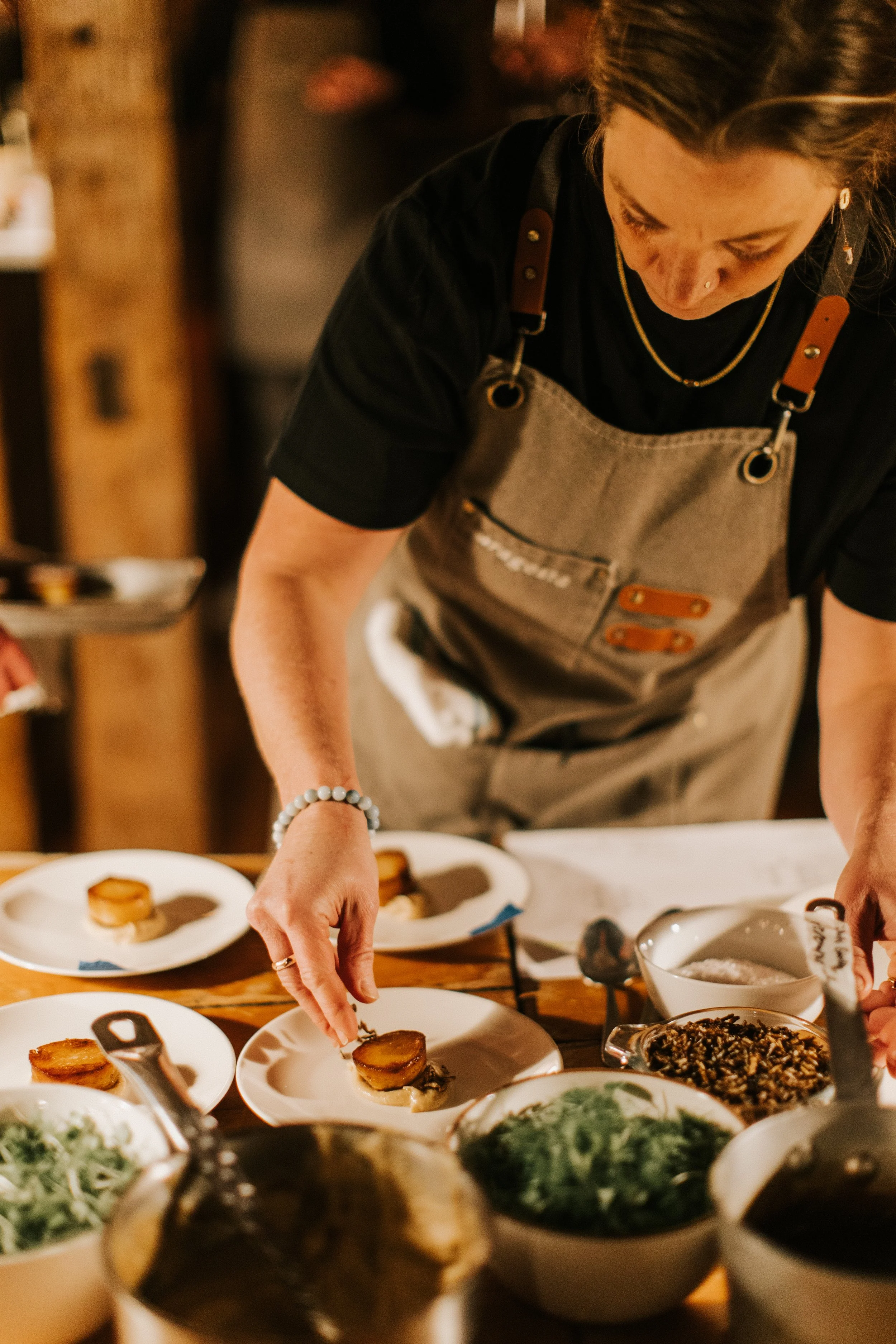 A woman prepares plated food at a kitchen or restaurant counter, with various dishes and ingredients around her.